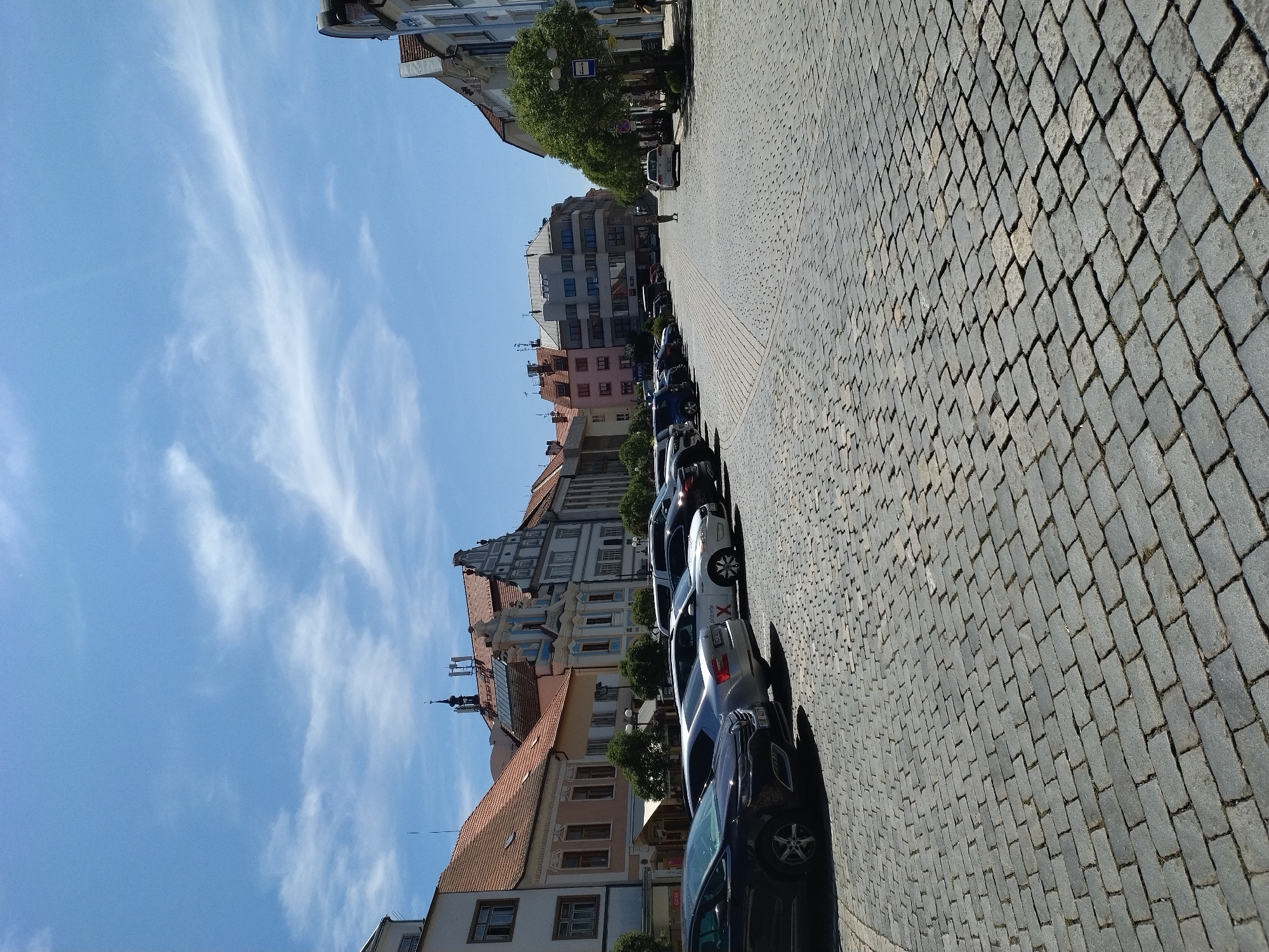 The image depicts a cobblestone street in Písek, Czechia, likely the main square. The street is lined with parked cars on the left and buildings with traditional architecture on the right. The sky is clear blue with scattered white clouds. Several people are visible in the distance, walking along the street and sidewalks. The scene is set during daytime, under sunny conditions. The perspective is from ground level, looking down the street towards the horizon.
