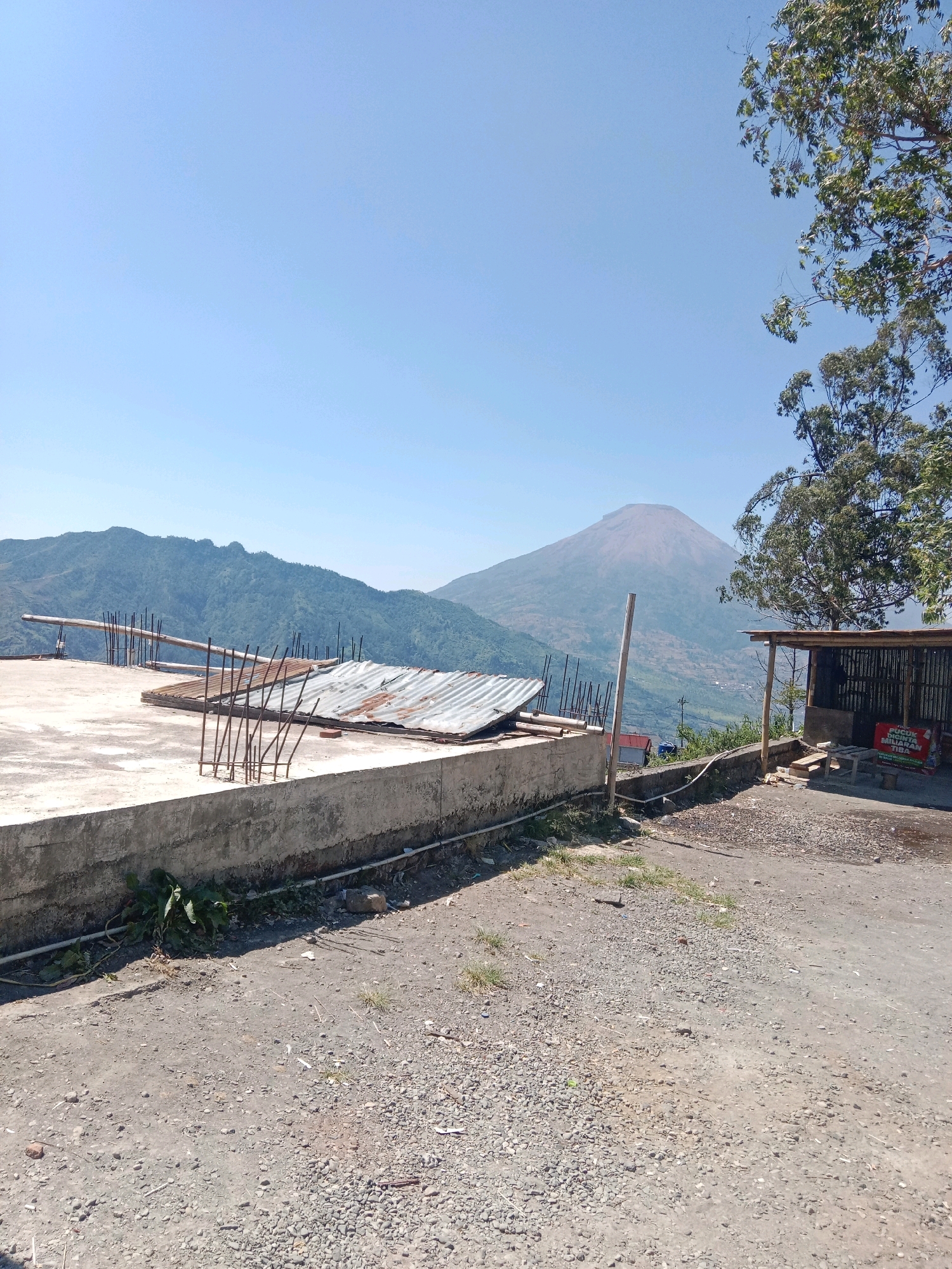 The image captures a scenic view of a mountain in the distance, likely Mount Prau, located near Tieng, Indonesia. In the foreground, a concrete platform is visible, part of an unfinished construction. Several rusted metal rods protrude from the concrete, and a sheet of corrugated metal rests on top. To the right, a small wooden structure with a thatched roof serves as a roadside stall. A sign with Indonesian text is affixed to the stall. The ground in front of the stall is covered with gravel and dirt. Lush green trees frame the right side of the image, with their branches reaching towards the clear blue sky. The background is dominated by rolling hills and the majestic mountain, which appears to be a dormant volcano. The overall impression is one of a rural, mountainous landscape with evidence of ongoing development.