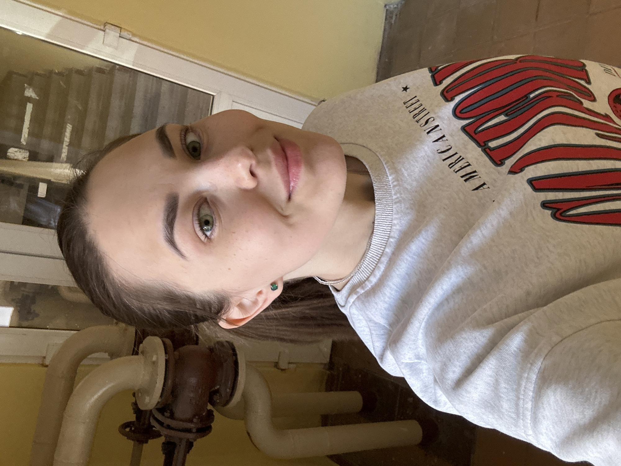 A medium shot photograph shows a fair-skinned young woman with brown hair and green eyes indoors, likely in Perm, Russia, based on visual cues such as building architecture. She is wearing a light gray sweatshirt with a red and blue design and text that reads "AMERICAN STREET." She is facing the camera with a slight smile. Visible behind her are pipes, a wall, and possibly a window frame. The lighting in the room appears to be natural. The overall environment suggests a building interior in Perm, Russia.