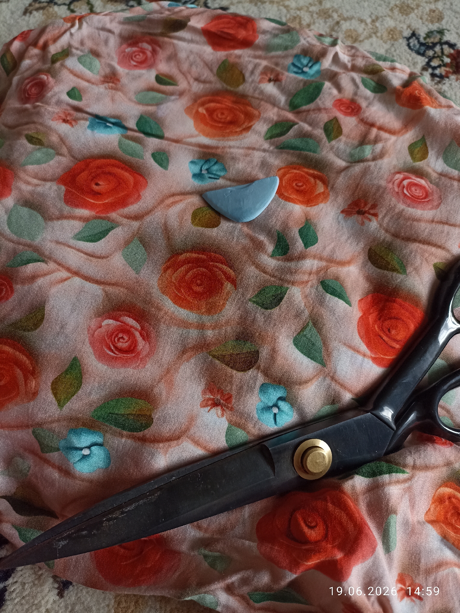 The image displays a close-up, overhead view of a pink fabric with a floral pattern of red and orange roses and scattered blue flowers and green leaves. A light blue tailor's chalk is placed on the fabric, just above and to the left of a large pair of black-handled scissors. The scissors are positioned diagonally across the lower right portion of the fabric, with their blades partially visible. The fabric is draped and wrinkled, suggesting it might be laid out for cutting. The background is a blurred carpet pattern in shades of beige and brown. A timestamp "19.06.2026 14:59" is overlaid on the bottom right corner of the image. No people or specific urban environmental cues are present.