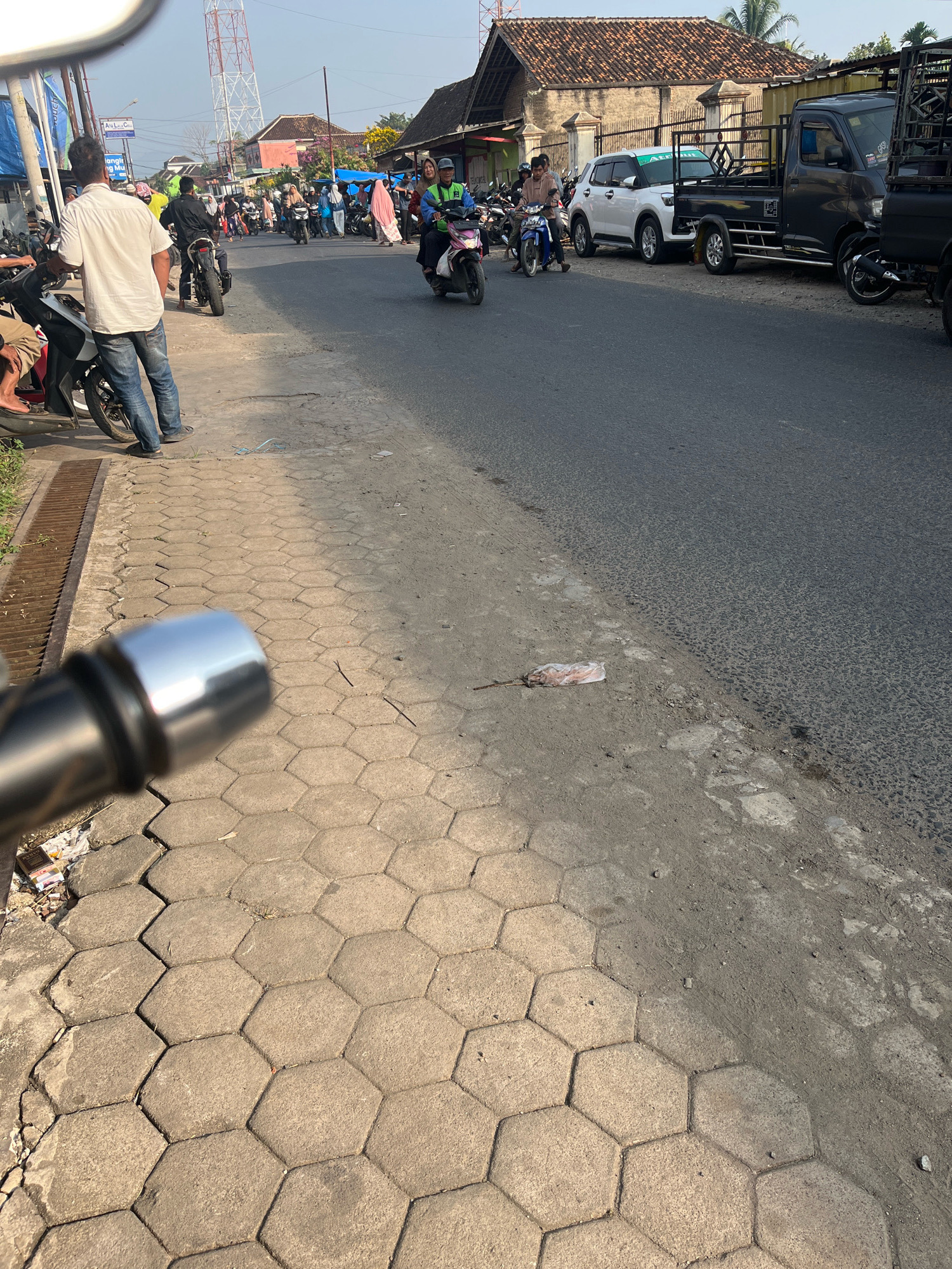 The image depicts a street scene in Kemuning, Indonesia. The foreground features a paved sidewalk made of hexagonal concrete blocks, with a metal drain cover along the left edge. A motorcycle's chrome handlebar is visible in the bottom left. The street itself is asphalt, with some dirt and debris on the shoulder. Several people are present, mostly on motorcycles. On the left side of the street, a man in a white shirt and blue jeans stands near a parked motorcycle. Further down the street, a procession of motorcycles and riders is visible, some with passengers. Vehicles are parked along the right side of the street, including a white SUV and two dark pickup trucks. Buildings with tile roofs line the right side of the road, with some vegetation and a tall tower in the background. The lighting suggests it is daytime, likely late morning or afternoon given the shadows. The overall impression is of a busy, active street with vehicular and pedestrian traffic.
