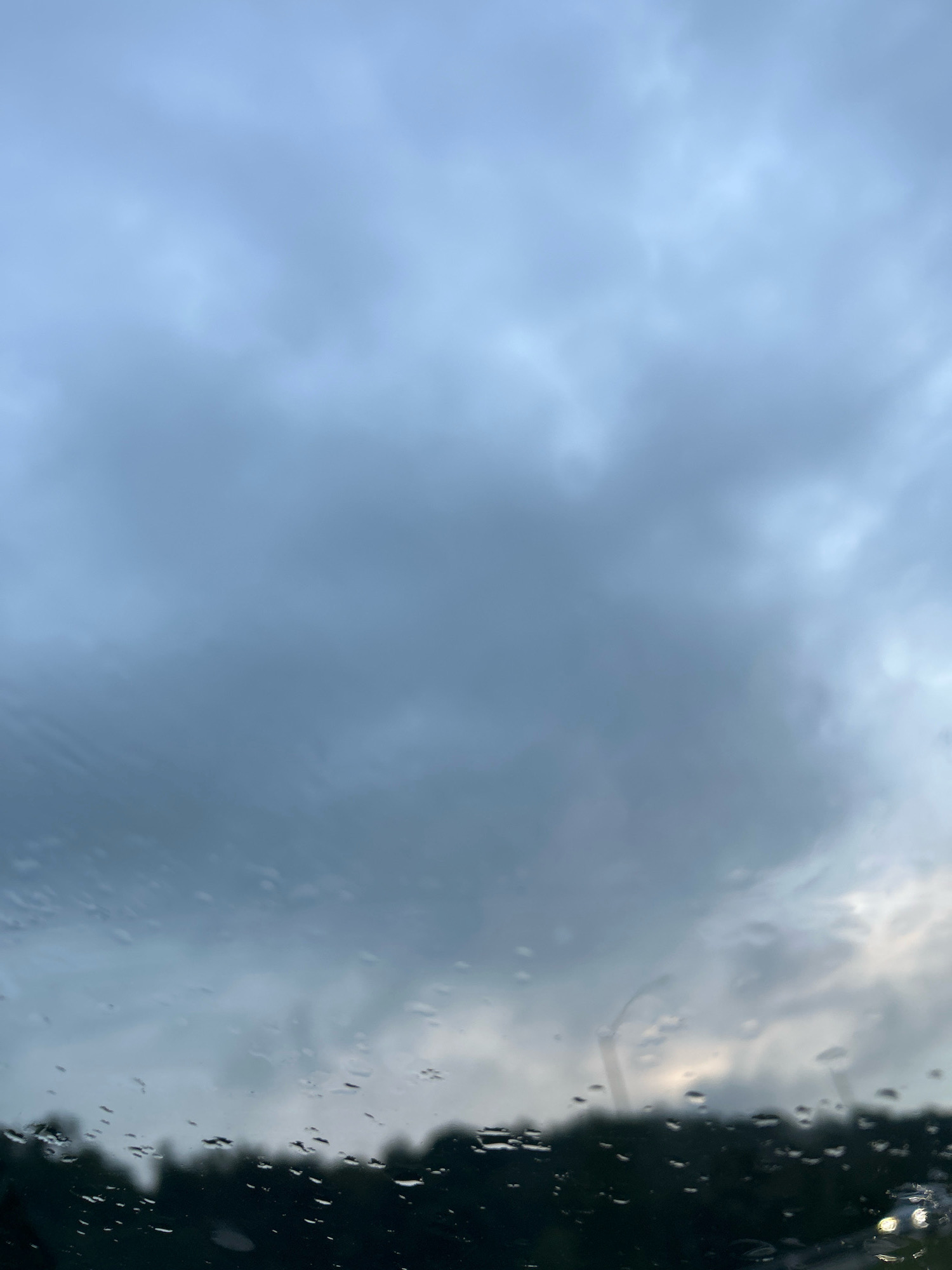 A close-up, low-angle shot captures a cloudy sky through a window with raindrops. The sky is filled with various shades of gray and blue clouds, with some lighter areas suggesting diffused sunlight. In the foreground, numerous water droplets are visible on the glass, distorting the view of the blurry, dark silhouette of trees and a faint hint of streetlights or car headlights in the bottom right corner. The scene suggests a rainy day in Robinson Township, USA.