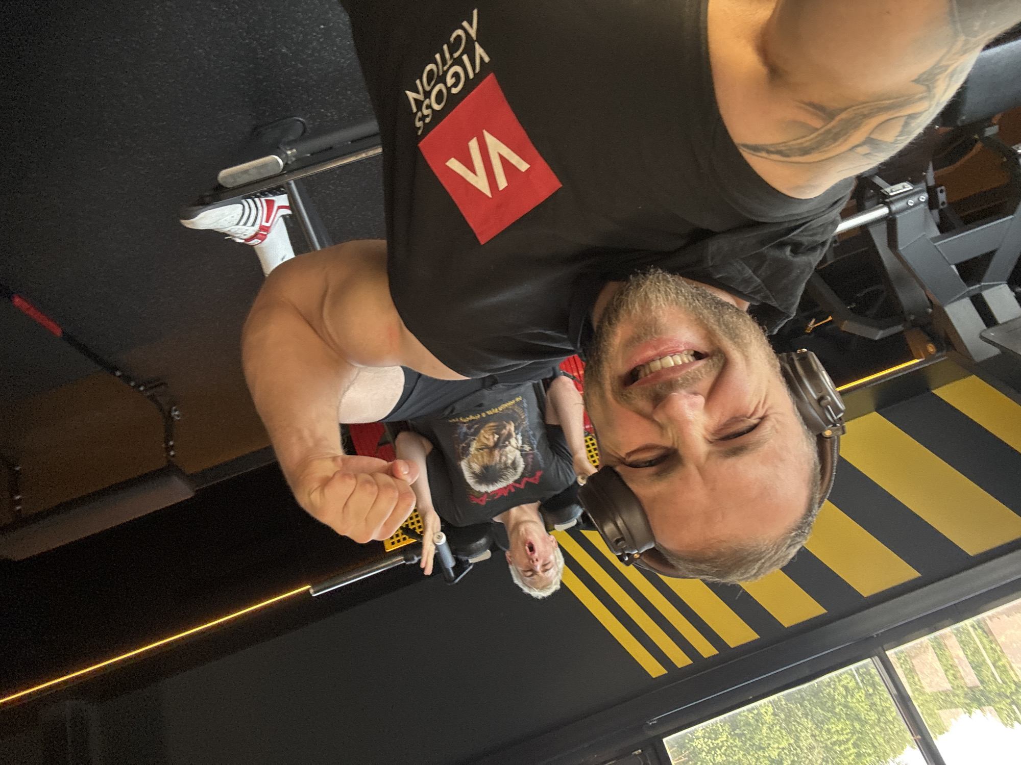 Two men are in a gym. The man in the foreground, on the left, is taking a selfie. He has headphones on, a beard, and is wearing a black sleeveless shirt with a red logo that says "VA" and "VIGOSS ACTION". He has a tattoo on his left arm. He is smiling and winking. The second man is in the background, on a weight machine, appearing to be exercising. He has blonde hair and is wearing a black t-shirt with a picture of a man on it. The gym has black walls with yellow diagonal stripes. There is a window in the upper left corner showing greenery outside.