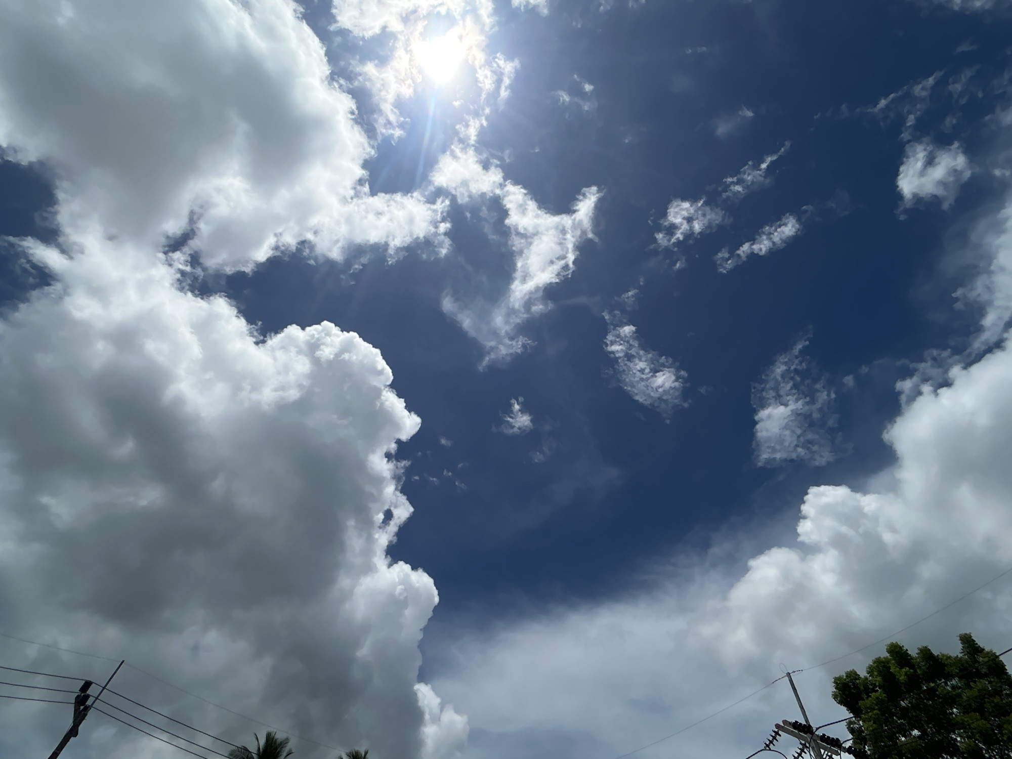 The image shows a view of the sky in Baan Hin Kong, Thailand. The sky is a deep blue, with scattered white clouds. The sun is shining brightly, creating a glare and sunbeams in the upper center of the frame. There are utility poles and wires visible in the lower left and lower right corners, with some greenery from a tree in the lower right.