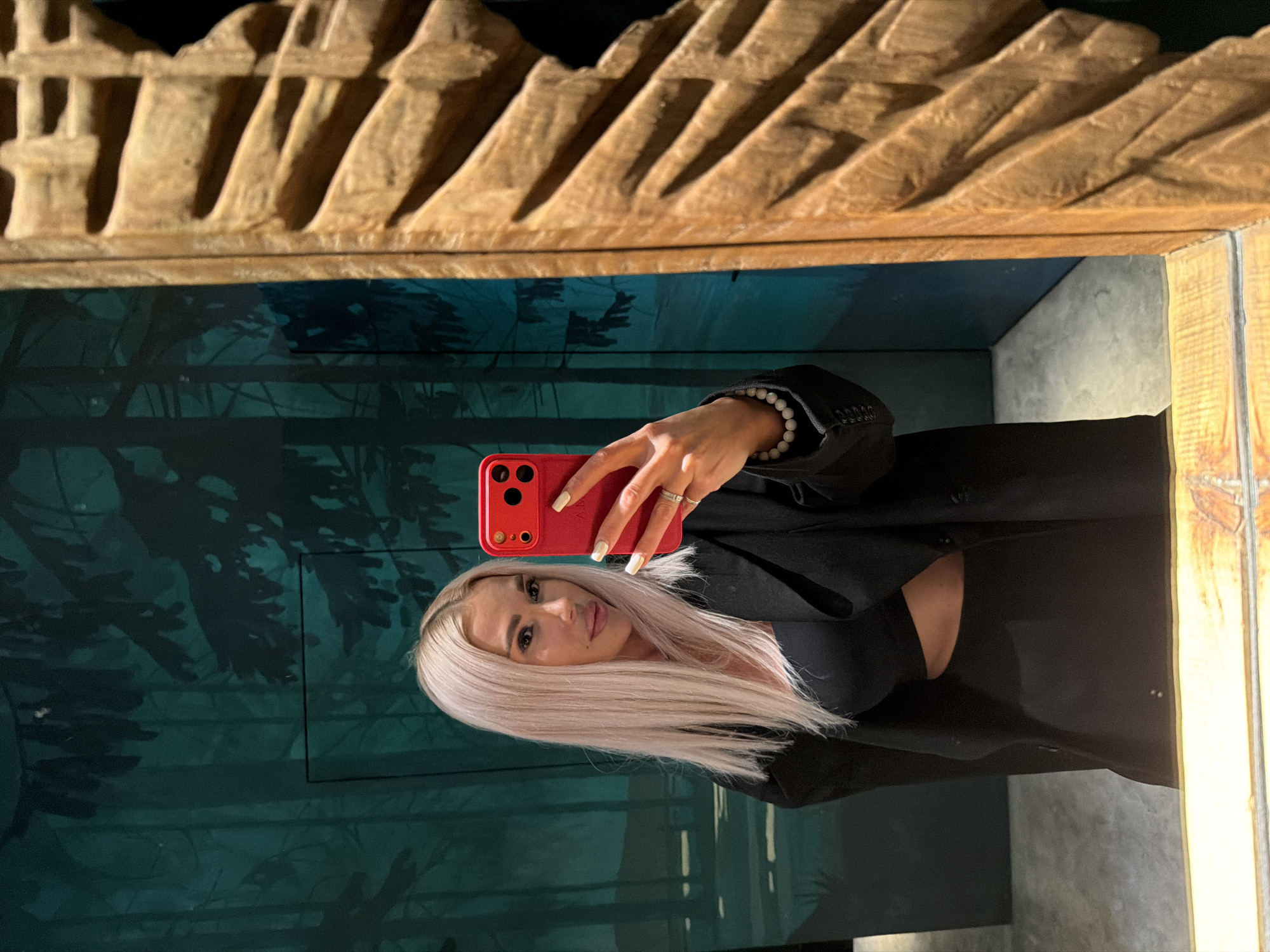 A woman with long, straight light blonde hair is captured in a mirror selfie. She has fair skin and a neutral expression, looking directly at the camera. She is wearing a black top and a black blazer or jacket. On her right wrist, she wears a white beaded bracelet, and rings are visible on her fingers. She holds a red smartphone, which has two visible camera lenses, in her right hand to take the photo. Her fingernails are painted white.
The mirror is framed by an ornate, carved wooden border at the top and partially on the right side. The light-colored wooden frame features a textured, possibly mountainous or abstract peak-like design.
The background reflected in the mirror behind the woman is a deep teal or green wall, adorned with dark, silhouetted patterns resembling trees or foliage, creating an artistic or themed ambiance. The lighting appears artificial, highlighting details like the wooden frame's texture. The scene suggests an indoor location, possibly an art space or a specially decorated room. There is no visible text in the image.