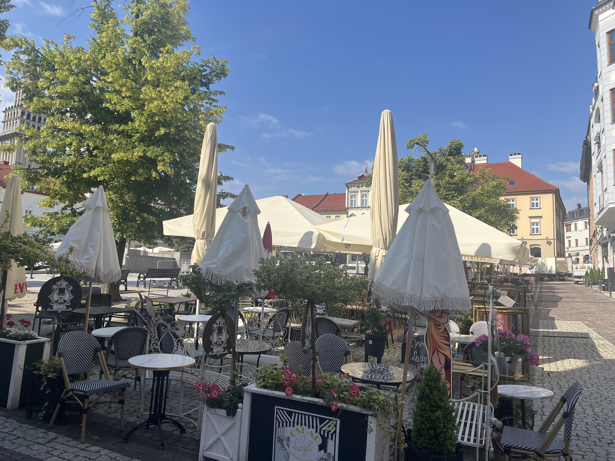 This is a ground-level, medium-shot, outdoor photograph of a cafe seating area in an urban setting. The cafe is set up on a cobblestone street with several tables and chairs arranged beneath large, closed umbrellas. The umbrellas are predominantly white and cream colored. The tables vary in size and design, with some being round and white-topped, while others appear to have darker, patterned surfaces. The chairs are a mix of styles, including some with black and white woven seats and backs, and others with more intricate metal designs.
In the foreground, several planters contain green foliage and pink flowers. One planter features a sign that reads "OSMAN Cafe". A life-sized cardboard cutout of a woman in an orange dress stands next to a small table.
In the background, there are more tables and chairs, suggesting a larger seating area. Beyond the cafe, buildings with red-tiled roofs and light-colored facades are visible, indicating a European architectural style. A tall, ornate tower with a pointed roof rises in the left background. Trees with green foliage are scattered throughout the scene, providing shade. The sky is clear and blue. The overall impression is of a quaint and inviting outdoor dining space.