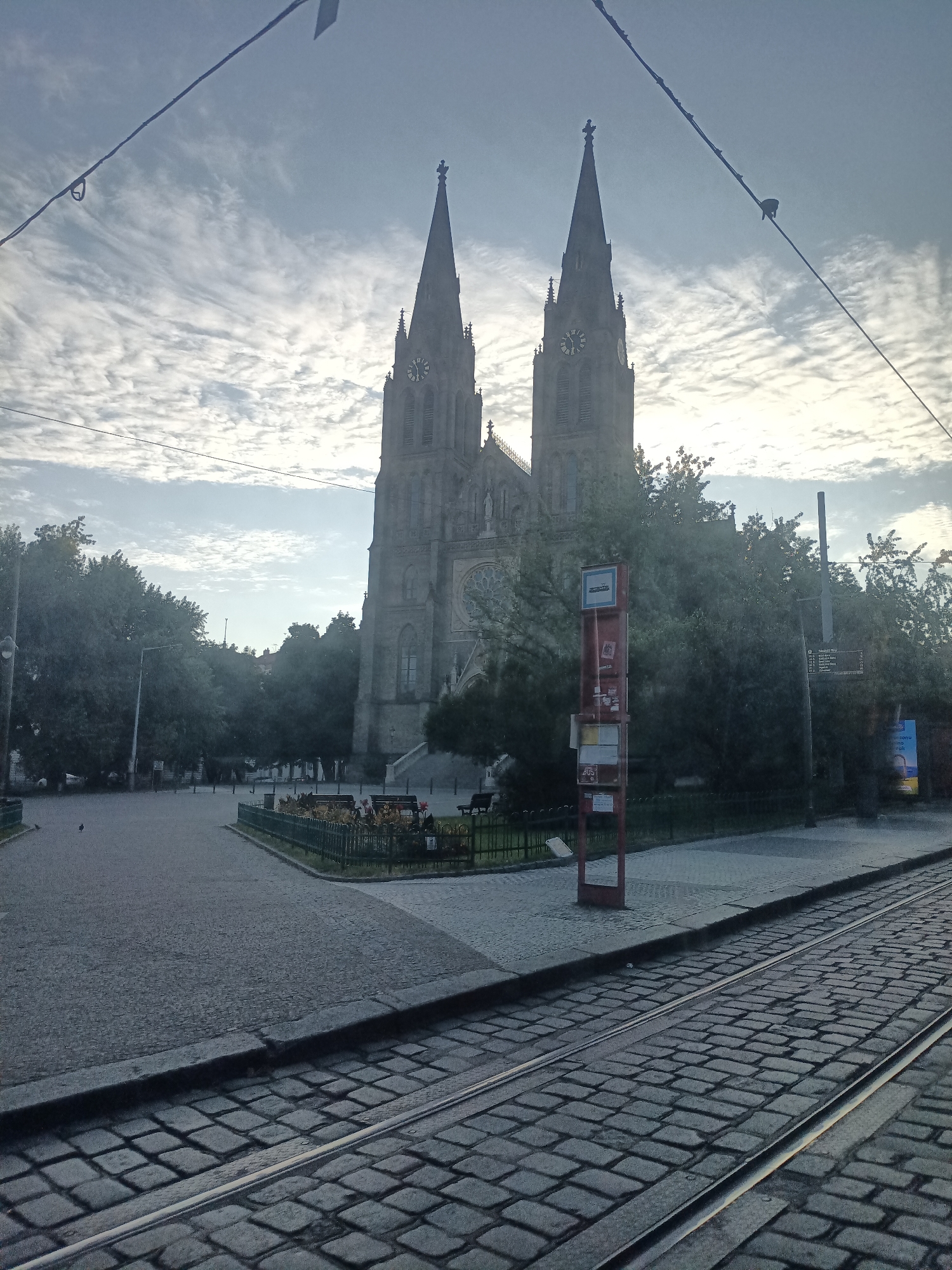 A view of the Basilica of St. Peter and St. Paul in Prague, Czechia, as seen from a tram. The basilica, with its twin spires and clock faces, dominates the background under a cloudy sky. In the foreground, cobblestone tracks for a tram are visible, suggesting the photo was taken from inside a moving tram. A red bus stop shelter with information signs stands to the right of the basilica, surrounded by trees and a paved walkway. The area appears to be a public square or park, with benches and a low fence lining a grassy area.