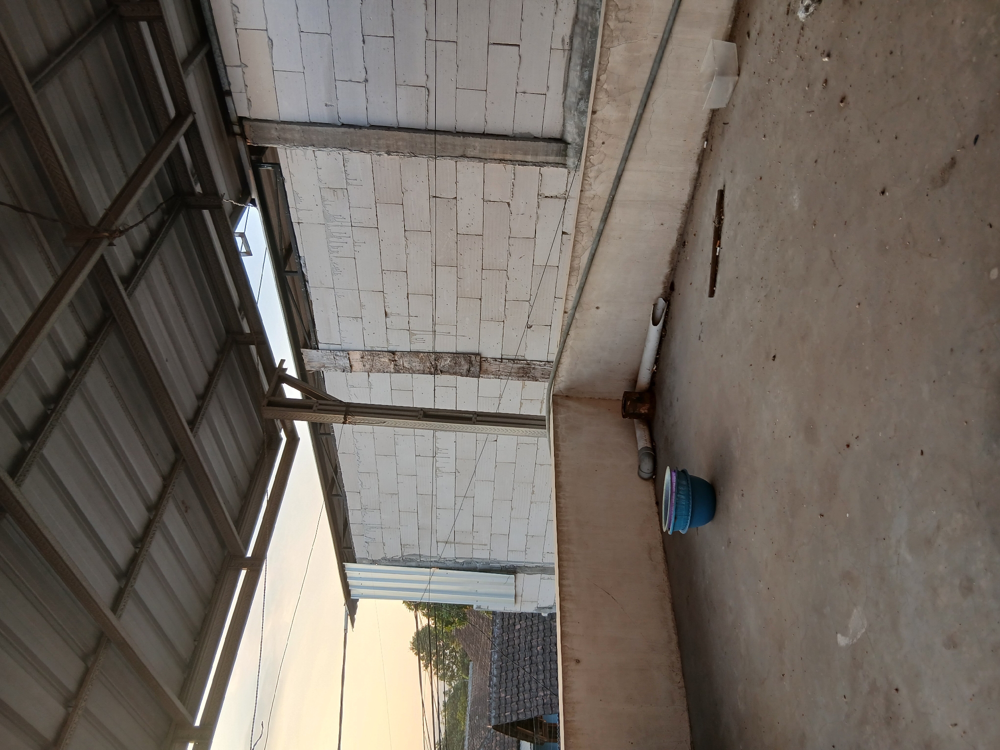 An exterior shot captures a concrete rooftop area with a corrugated metal roof supported by rusted metal beams and wooden poles. The wall behind the rooftop is constructed of white cinder blocks, with a section covered by corrugated metal siding on the left. The rooftop is littered with various objects, including two blue plastic basins stacked on top of each other, a length of white pipe with an opening, a rusty cylindrical object, a piece of wood, and a clear plastic container. In the background, a residential neighborhood with tiled roofs and trees is visible under a pale sky. The scene is located in Kademangan Kulon, Indonesia.