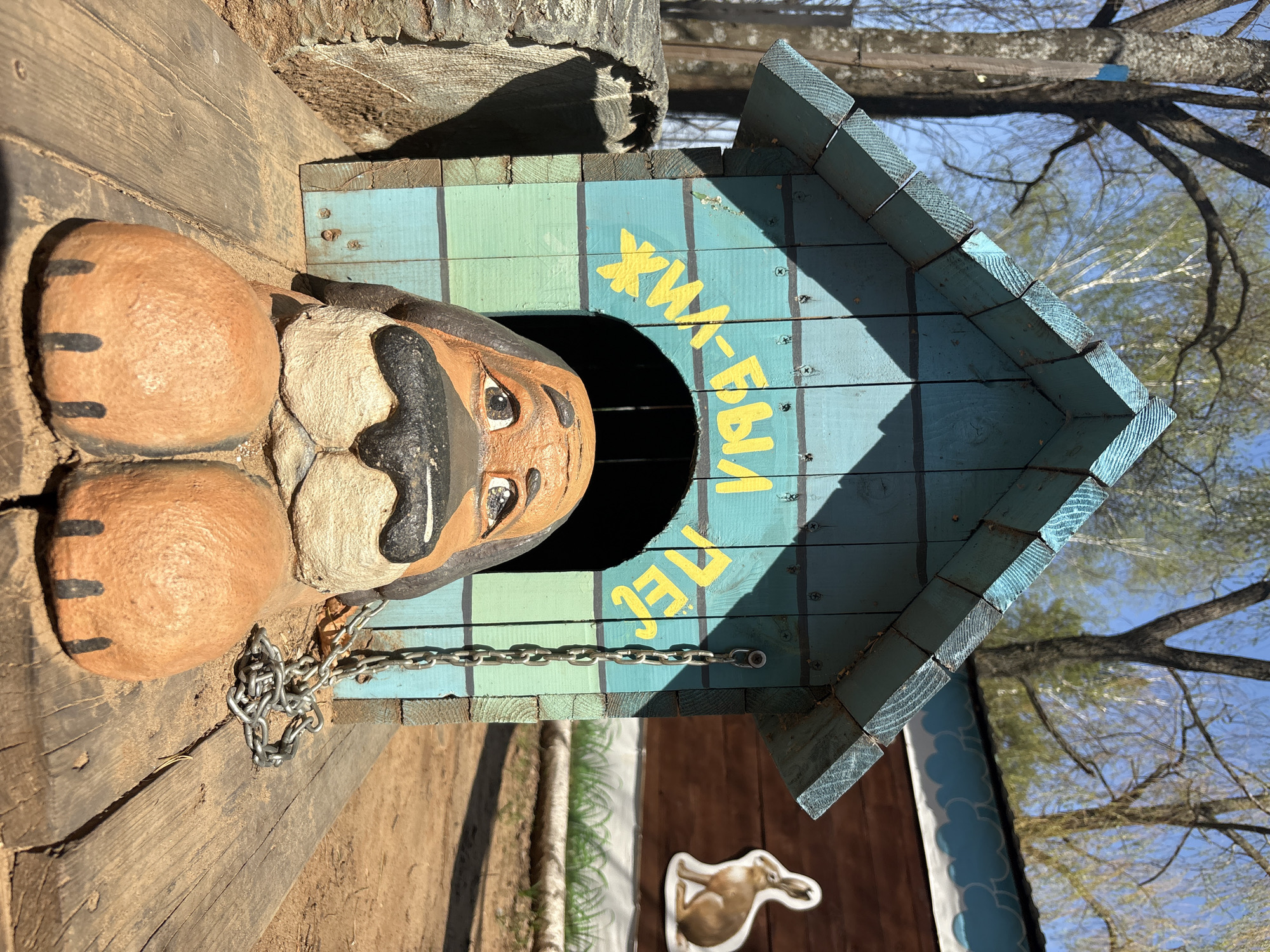 A dog sculpture is inside a blue and green doghouse. The dog appears to be made of a tan material and has black markings. The doghouse is located outside in the city of Chaikovsky, Russia. The chain appears to be attached to the dog.