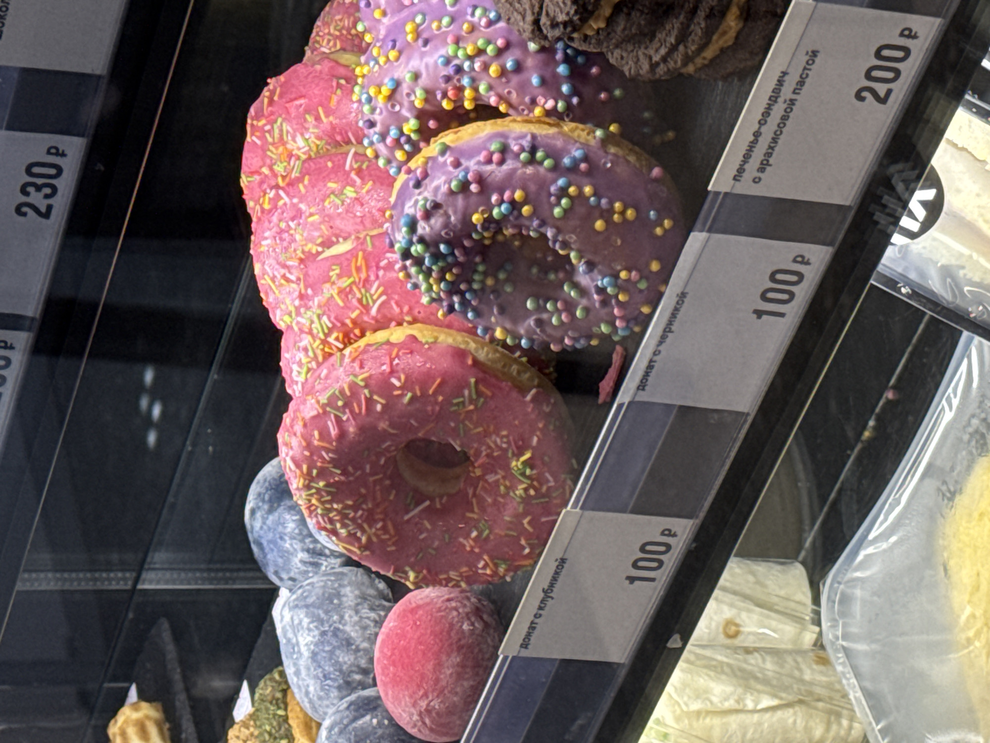 This image captures a close-up, slightly tilted view of a dessert display case, likely in a bakery or supermarket in Moscow, Russia. The primary subjects are a vibrant assortment of donuts and other sweet treats.
In the foreground and center, several donuts are stacked. Two are prominently featured with bright pink icing and colorful sprinkles. Another two have purple icing with similar multi-colored sprinkles. One of the purple donuts shows a bite taken out of it. To the right, partially visible, is a dark brown, possibly chocolate, cookie sandwich or donut.
Below the pink donuts, a stack of blue/grey spherical items (possibly mochi or another confection) is visible, along with a single red spherical item. The display case has glass shelves and reflective surfaces.
Several price tags with Cyrillic text and prices in Russian Rubles (₽) are attached to the front of the shelves. Visible text includes:
* "донат с клубникой" (donut with strawberry) priced at "100 ₽".
* "донат с черникой" (donut with blueberry) also priced at "100 ₽".
* "печенье-сэндвич с арахисовой пастой" (cookie-sandwich with peanut paste) priced at "200 ₽".
* Another price tag shows "230 ₽", likely for an item above or to the left that is mostly out of frame.
The overall scene suggests a retail environment showcasing sweet baked goods and confections for sale. The colorful nature of the donuts makes them visually appealing.