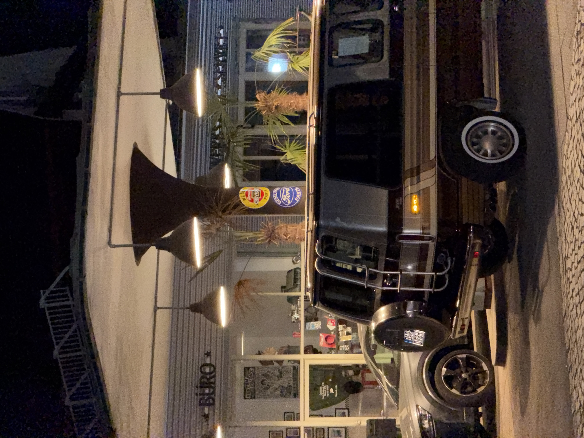 The image shows a dark, exterior view at night, possibly a gas station or a covered outdoor area. A vintage dark brown and tan van with a spare tire mounted on the rear is parked in the foreground. A silver car is parked to the left of the van. In the background, there is a building with a striped facade, large windows, and signs that appear to read "BURO". Decorative pendant lights hang from the ceiling structure. Several potted palm trees are placed around the area. A circular sign with the Chevrolet logo and text "GENUINE PARTS" is visible in one of the windows. Another circular sign with the Ford logo and text "GENUINE PARTS" is also present. The ground is paved with cobblestones. The overall scene suggests a commercial establishment, possibly catering to automotive enthusiasts or a retro-themed business. There is no specific indication of the location being in Garmisch-Partenkirchen, Germany.