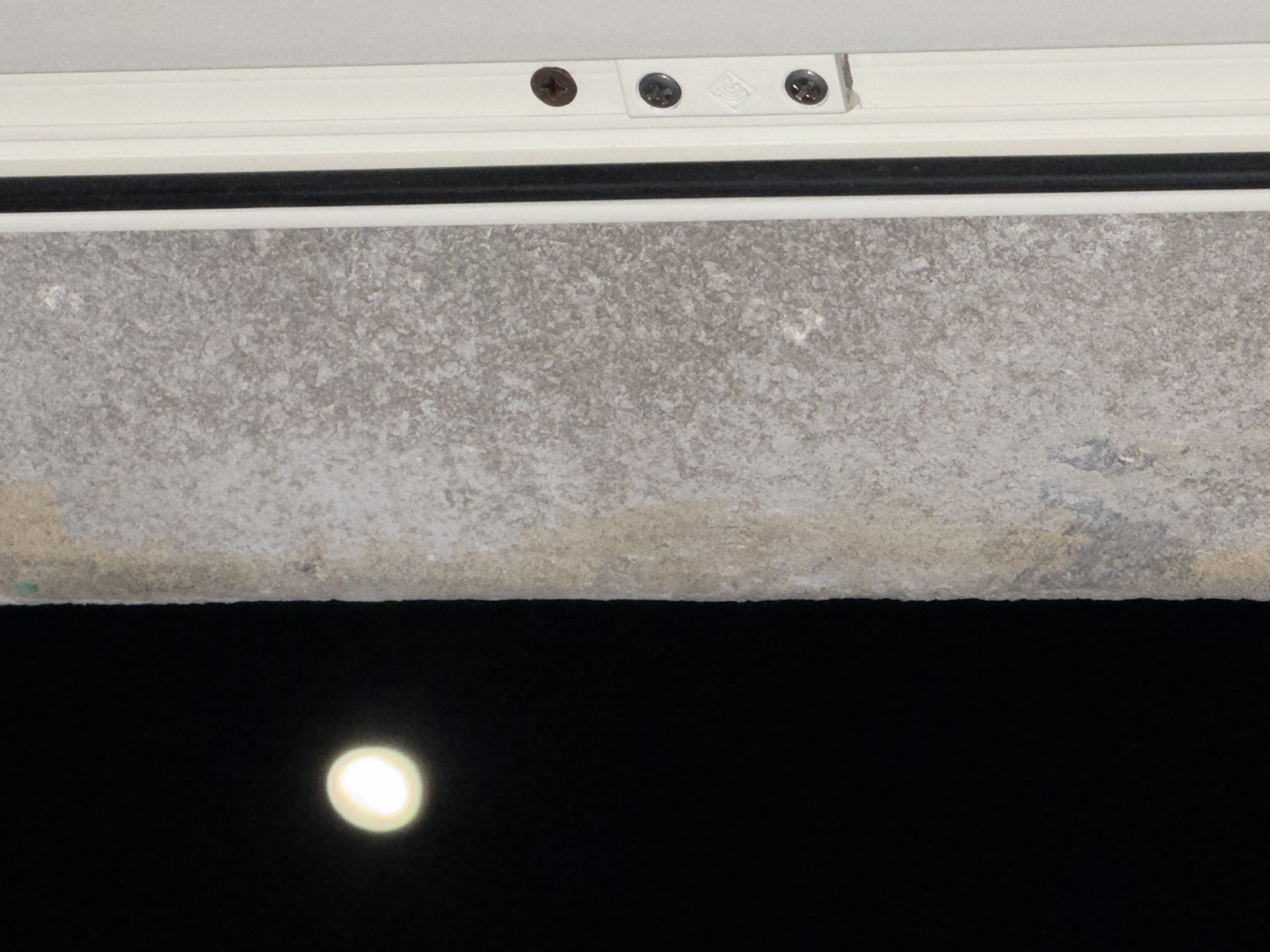 The image shows a close-up view of a windowsill with a white frame secured by screws. Below the windowsill is a concrete ledge, showing signs of wear and discoloration. The background is a dark night sky with a blurred bright light source, likely the moon. There is no specific information indicating the location in Catania, Italy, or any people, actions, or interactions.