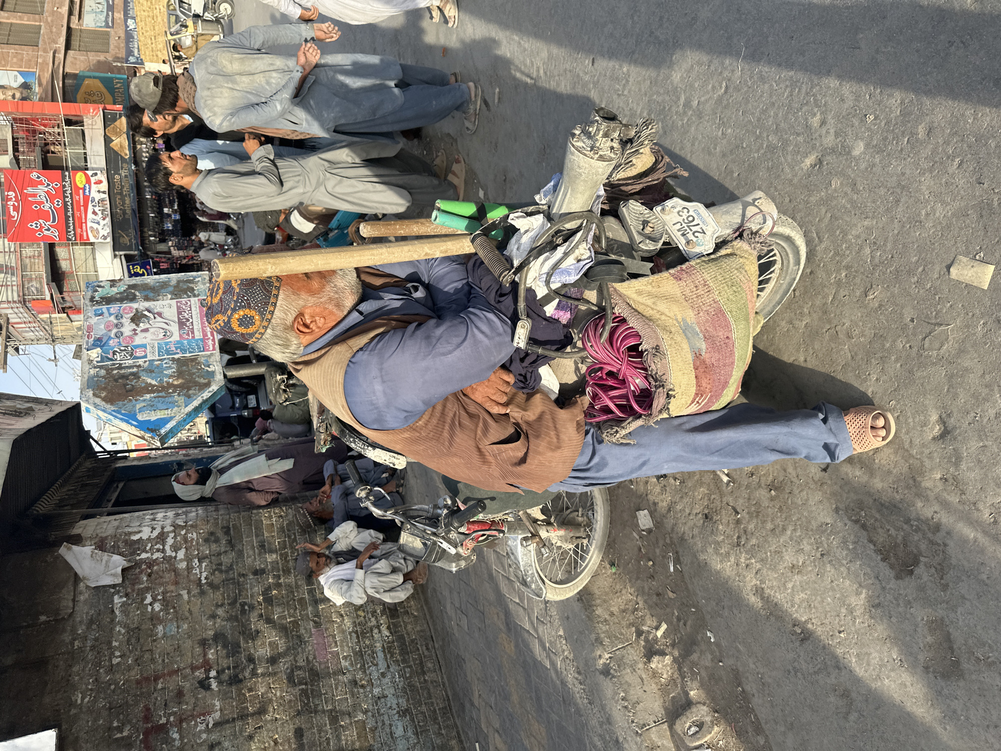 The image captures a street scene in Quetta City Tehsil, Pakistan. In the foreground, an elderly man with a beard and wearing a traditional embroidered cap, a blue shirt, and a brown vest leans on a motorcycle. He is also wearing blue pants and light-colored sandals. The motorcycle has a license plate that reads "MJ 2763." On the back of the motorcycle, a woven bag is attached, containing bright pink coiled rope and other items.
Behind the man and the motorcycle, several other individuals are visible. To the left, two men sit on the ground next to a brick wall. One, wearing a white shalwar kameez, gestures with his hand. Another man, dressed in grey, is also seated. Further back, a man in a light-colored headscarf stands next to a doorway.
The environment appears to be a bustling urban street with shops and signage. A red banner with Arabic text is prominent in the background. The ground is paved with bricks and shows signs of dust and debris. The overall lighting suggests it is daytime, with shadows cast by the structures and individuals. The scene conveys a sense of everyday life and commerce in the area.