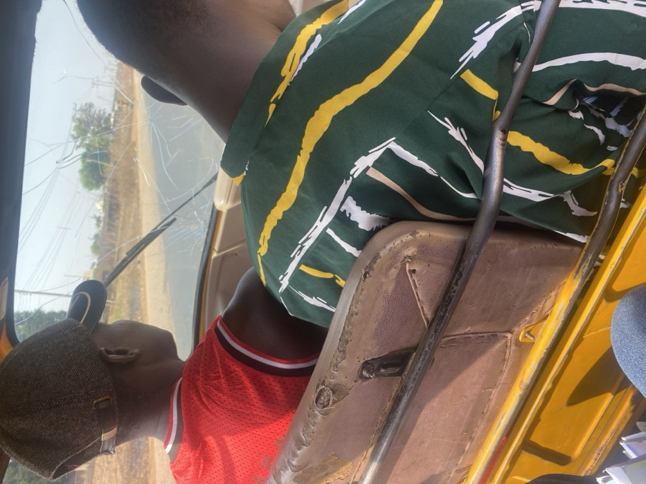 The image captures a scene inside a vehicle, likely a three-wheeled auto-rickshaw or similar utility vehicle, with two individuals present. The photo is taken from a slightly elevated, rear-facing perspective within the vehicle, looking forward.
In the foreground and center, a person is seated, presumably the driver, facing away from the camera. They are wearing a dark green shirt with prominent yellow, white, and light brown stripes. The fabric of the shirt appears somewhat creased. Their dark-skinned arm is visible on the left side of the frame. The seat they are on, partially visible below them, is upholstered in a worn, light brown or tan material, showing signs of aging and possibly damage, with some areas appearing cracked or discolored. A dark metal bar, possibly part of the vehicle's frame or handlebars, runs vertically in front of this seat.
Behind and to the left of the driver, the head and upper torso of a second person are visible. This individual has dark skin and is wearing a bright red sleeveless jersey, possibly a basketball jersey, with black and white trim around the neck and armholes. They are also wearing a dark grey or black cap. This person appears to be a passenger, seated behind the driver and looking forward.
The most striking detail is the vehicle's windshield, which is severely cracked. Numerous large cracks radiate across the glass, obstructing the view of the outside world. A windshield wiper is partially visible at the bottom edge of the cracked glass. Through the cracked windshield, a bright, sunny outdoor scene is visible, suggesting it is daytime with clear weather. A road or dirt path can be seen stretching into the distance, flanked by some sparse vegetation or trees. The exterior of the vehicle, particularly on the right side of the frame, shows parts of its bodywork in a bright yellow color. The general condition of the vehicle, particularly the cracked windshield and worn seats, suggests significant use or damage.