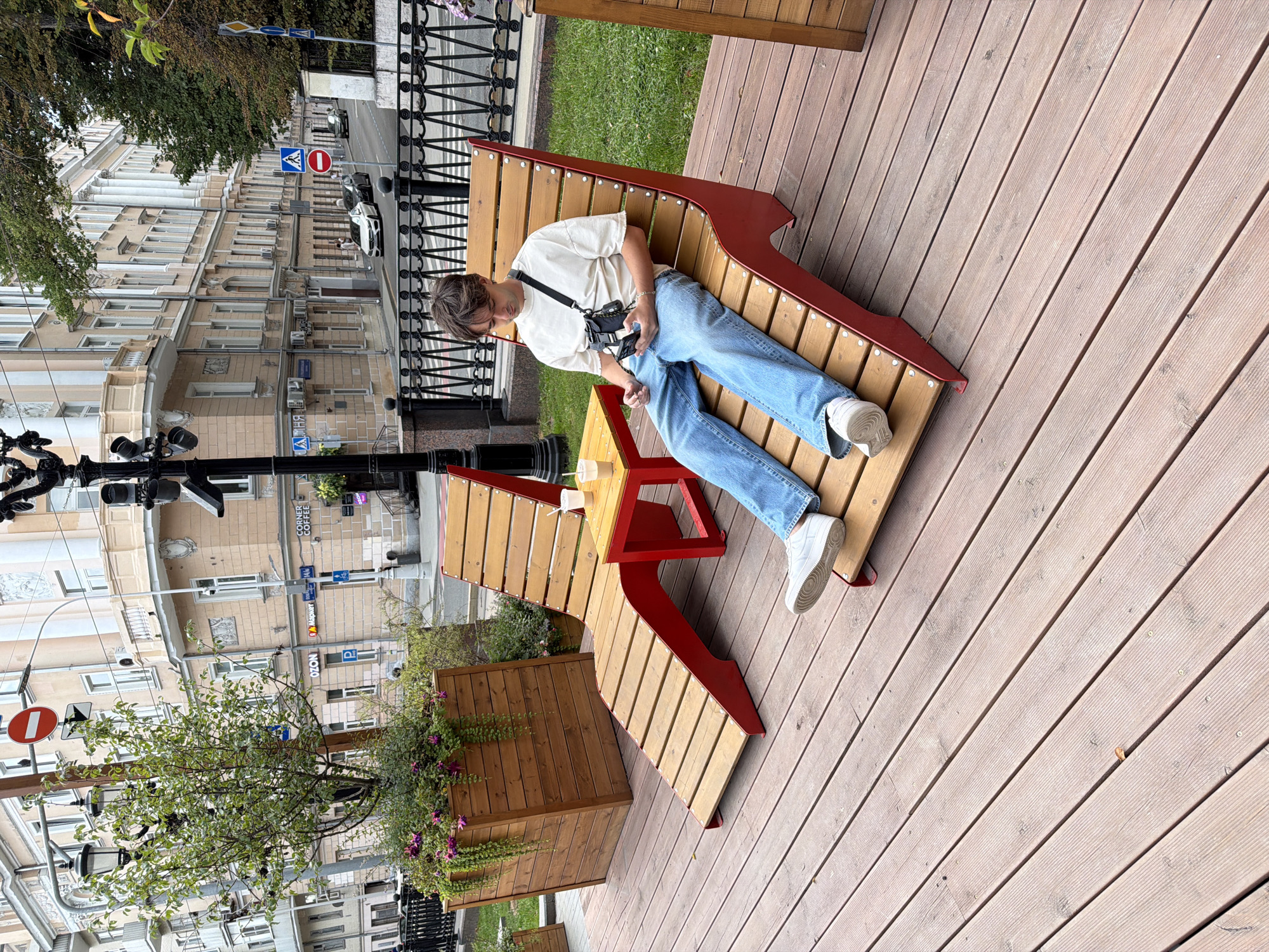 A young man sits on a red and wooden lounge chair outdoors, looking at his phone. He is wearing a loose-fitting beige t-shirt, blue jeans, and white sneakers. A black camera bag is slung across his body. He is sitting on a wooden deck in an outdoor seating area with another lounge chair and a small red table with two drinks on it. Behind him is a grassy area with a black metal fence and a tree in a wooden planter. In the background, there are buildings with a traditional European architectural style, indicating an urban environment. Traffic lights, road signs (including a "no entry" sign and a pedestrian crossing sign), and cars on a street suggest this is a public area in Moscow, Russia.