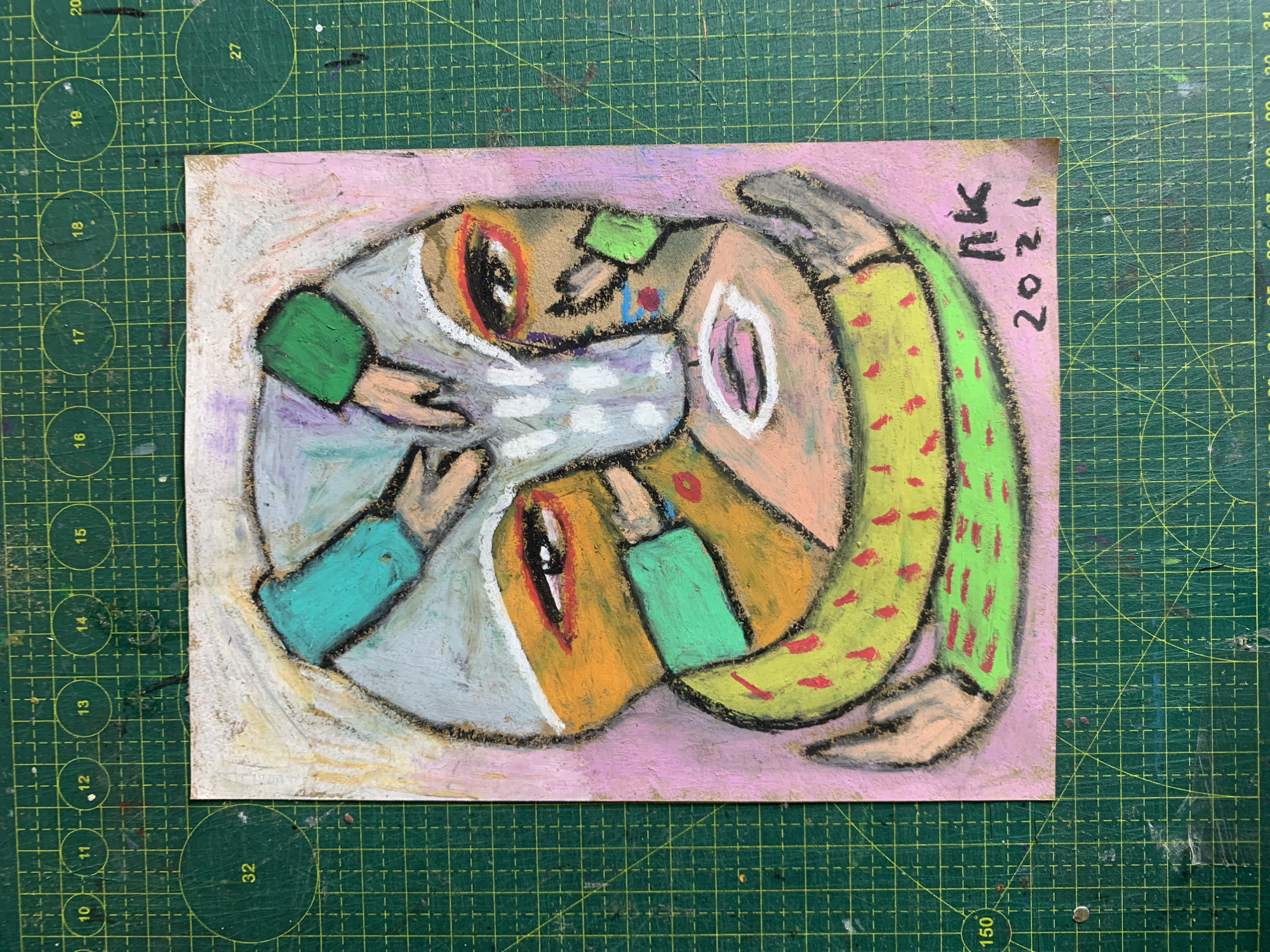 The image shows an abstract, colorful portrait drawn with pastels on paper. The drawing features a stylized face with exaggerated features, including large eyes, a prominent nose, and a gaping mouth. The face is surrounded by abstract shapes and lines in various colors, including green, orange, pink, and white. A signature "nk" and the year "2021" are visible in the lower right corner of the drawing. The drawing is placed on a green cutting mat with grid lines and numerical markings, which is commonly used for crafting and art projects. There is no indication or context provided within the image that places it in Sergeevo, Russia, or any other specific city.