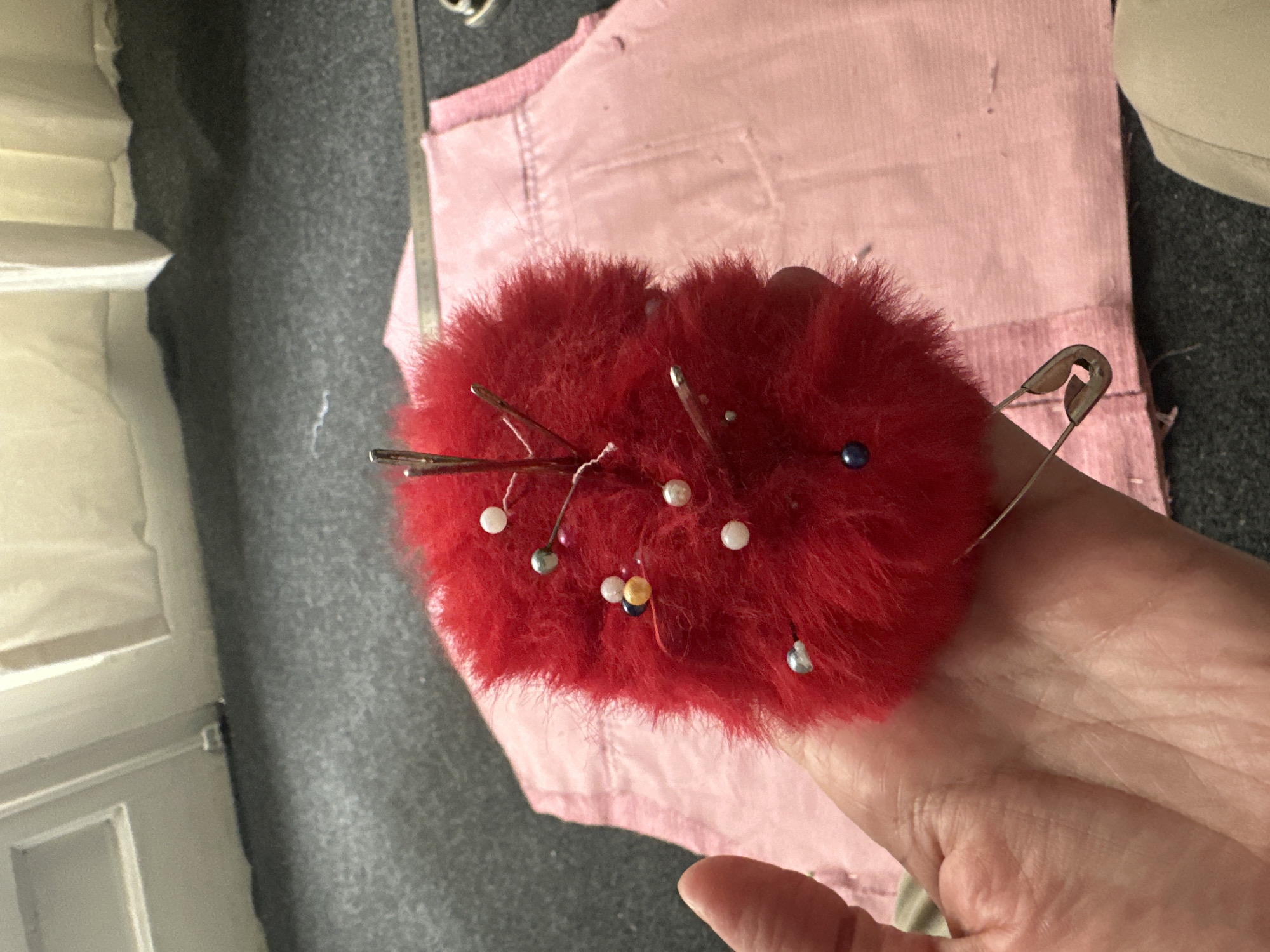 A hand holds a red, furry pincushion with several pins and a safety pin stuck in it. The background shows a pink fabric, a grey surface, a white cabinet or shelf unit, and a measuring tape. The location in Buenos Aires, Argentina is not discernible from the visual information.