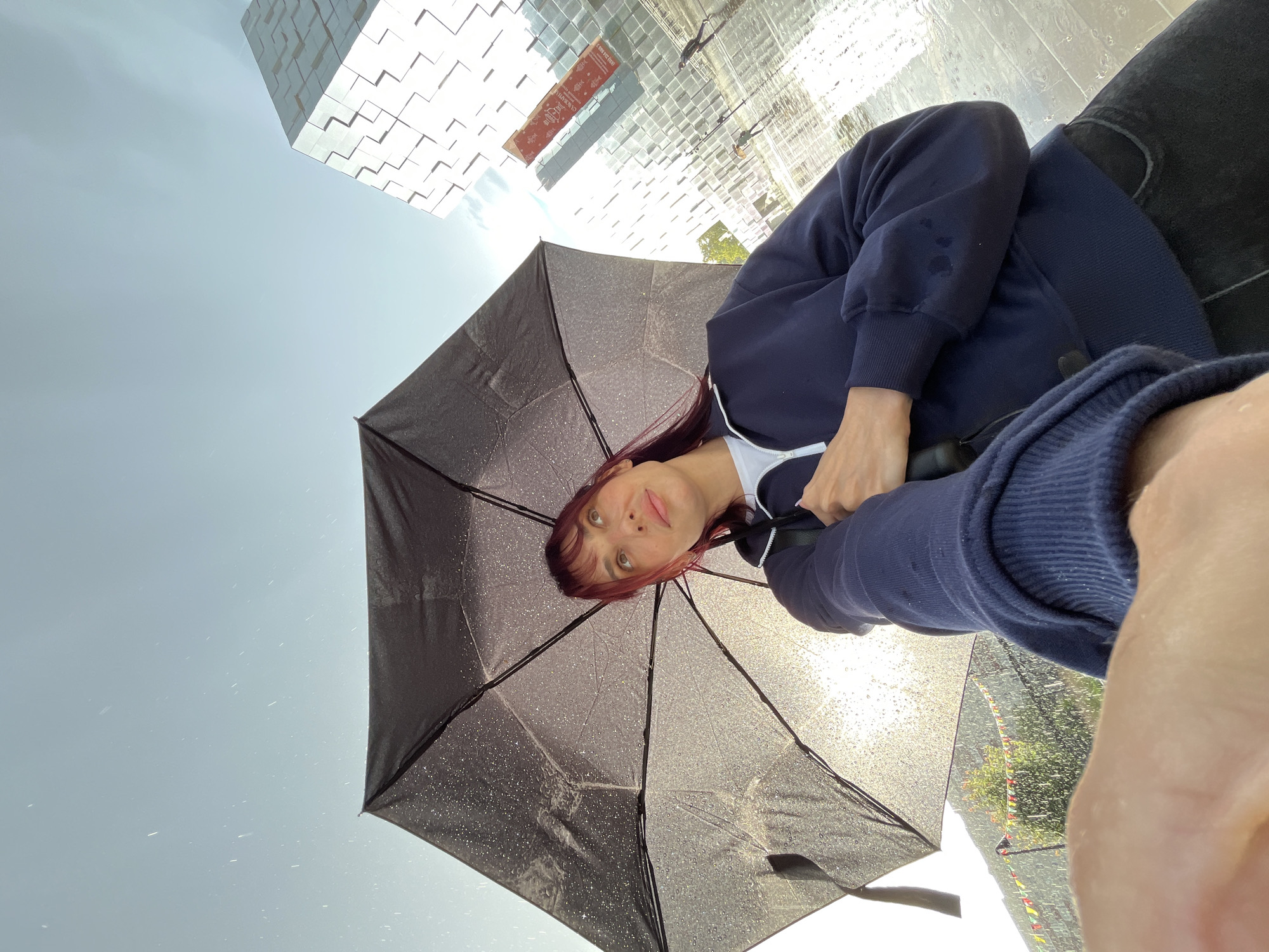 A low-angle, wide shot captures a woman holding a black umbrella from below as rain falls. She has dark, chin-length hair with red highlights and is wearing a navy blue zippered sweatshirt over a white shirt. The umbrella is speckled with raindrops, and sunlight glints through it, creating a slightly hazy effect. In the background, a modern, geometric building with a reflective, patterned facade dominates the right side of the frame. A red banner with Russian text hangs vertically on the building. The ground appears wet, reflecting the sky and building. Faintly visible in the distance are flags strung between poles and trees. The overall scene suggests an outdoor urban environment during a rain shower, possibly in Nizhny Novgorod, Russia, given the context of the question.