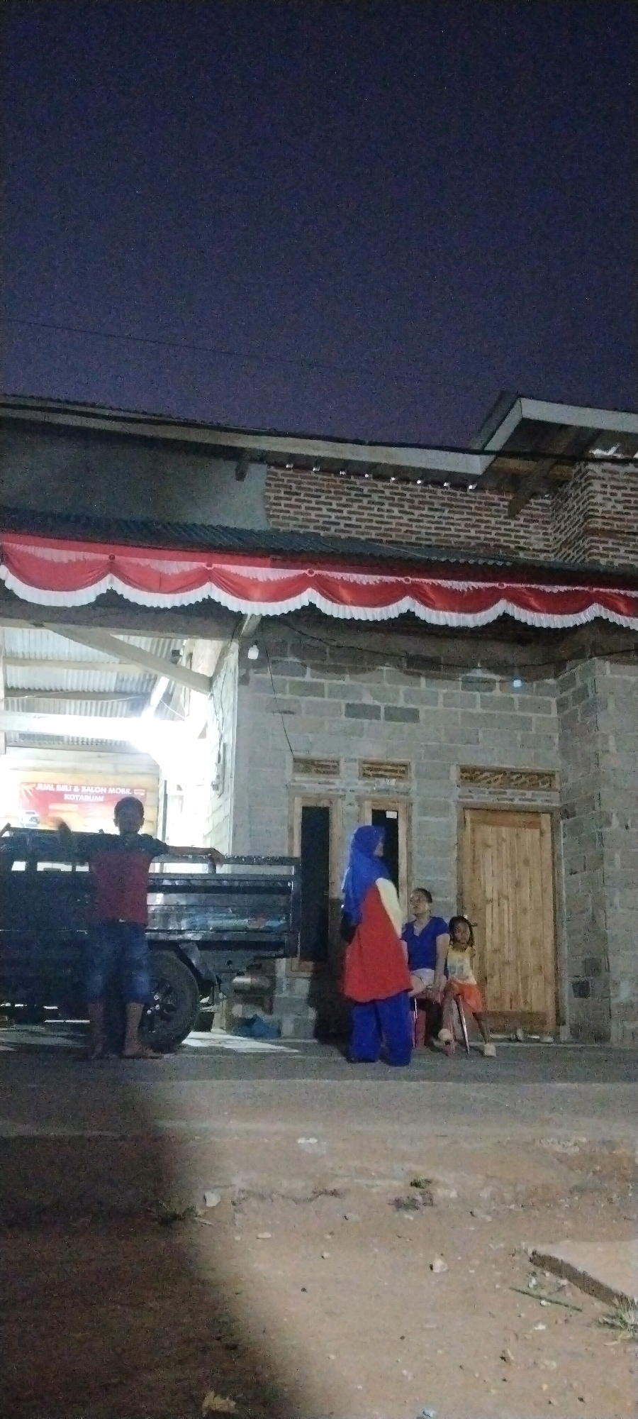 At night, on a dirt road in front of a building constructed from cinder blocks, a group of people are gathered. To the left, a man wearing a red shirt and blue shorts stands with his arms outstretched, leaning against the side of a black pickup truck. The truck has a metal bed and is parked on the road. In the center of the image, a woman in a blue hijab and a red and yellow dress stands near a doorway. To her right, a woman in a blue shirt sits on a chair next to a young girl wearing an orange shirt and shorts. The building behind them has a red and white banner draped across its facade, and a wooden door on the right. The sky above is dark.