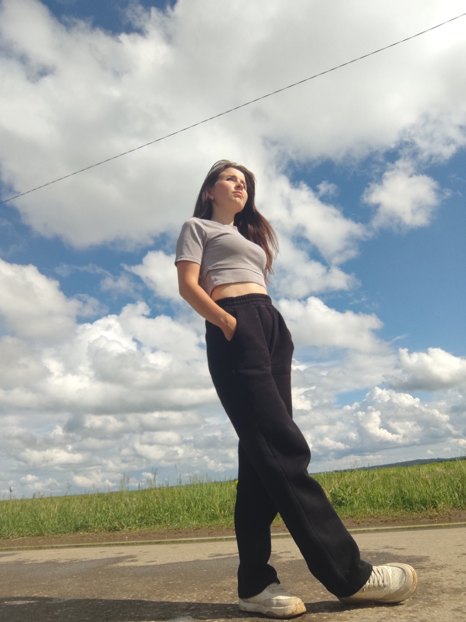 This is an outdoor, full-body portrait of a young woman standing on a paved road under a bright sky in Фоки, Russia. The woman, with long, dark brown hair, is looking upwards and to her left (viewer's right), exhibiting a serene or contemplative expression. She is dressed casually in a light grey short-sleeved crop top, black loose-fitting sweatpants, and white sneakers, which show some signs of wear. Her left hand is placed in her pocket, giving her a relaxed pose.
The background is dominated by a clear blue sky filled with large, fluffy white cumulus clouds, suggesting sunny and pleasant weather. A thin, dark horizontal line, possibly a utility cable, spans across the upper portion of the sky. Below the sky, a verdant green field stretches across the horizon, indicating a rural or natural setting. The foreground shows the asphalt road where the woman is standing, reflecting some light.
The scene takes place during the daytime, likely morning or afternoon, given the bright natural light illuminating the subject and clouds. No visible text is present in the image.