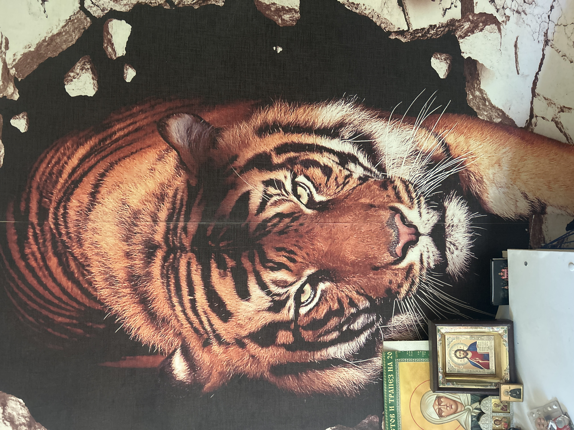 The image depicts a wall adorned with a large mural of a tiger emerging from a cracked wall. In the foreground, on a white surface, several religious icons are arranged. One is a larger icon of Jesus Christ, framed in dark wood. To its left is a smaller icon depicting a woman in a headscarf, and further left are two miniature icons. To the right of the Jesus icon, there is a small, dark item with a landscape painting of several buildings. There is also a green banner with Cyrillic text that appears to be a calendar or religious text. The wall behind the icons is covered by the tiger mural, which is the dominant feature of the image. The scene suggests a personal devotional space, possibly within a residential setting in Penza, Russia, given the presence of Russian text on the banner and the style of religious icons common in Orthodox Christianity.
