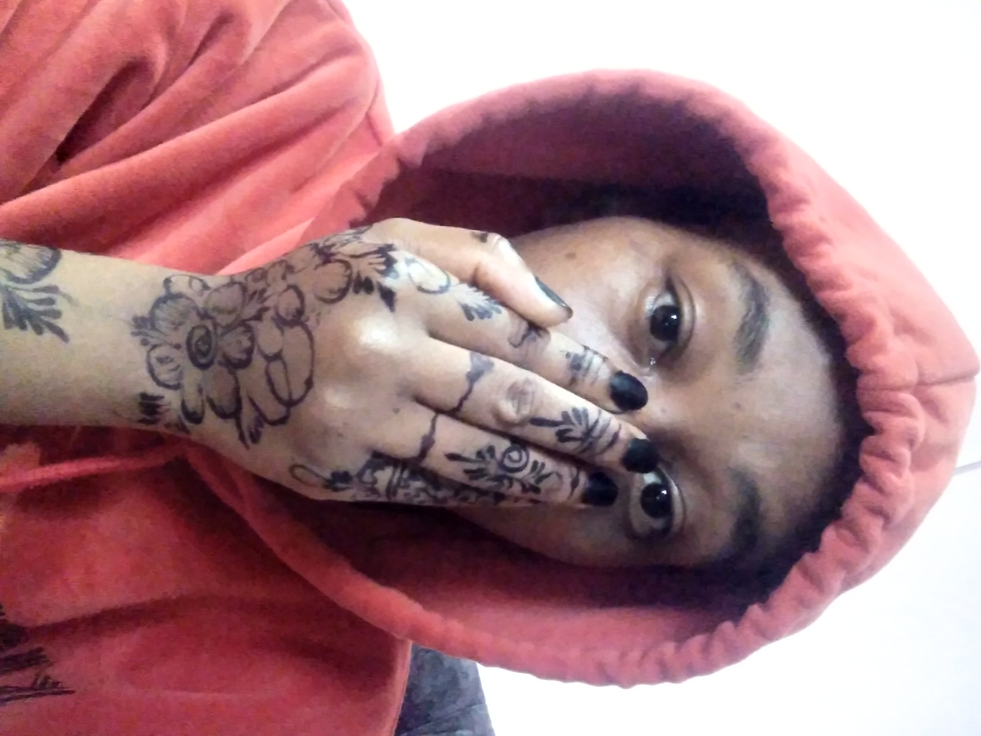A person with brown skin, wearing a reddish-orange hooded sweatshirt, is angled towards the right side of the frame. The hood is up, partially obscuring the top of the head. The person's right hand is raised, covering the lower half of the face, leaving the eyes and top portion of the nose visible. The fingernails are painted black. The hand and lower arm are adorned with black floral henna designs. The background is primarily off-white and out of focus. The city and specific location are unknown.