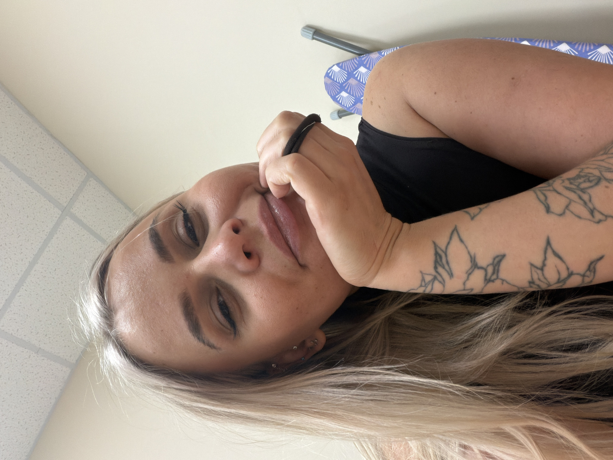 A woman is indoors, in a room with a suspended tile ceiling, a wall, and an ironing board, with the woman facing towards the camera. The woman is fair-skinned with light blonde hair. She is wearing a black tank top. The woman has tattoos on her right arm. She is holding a black hair tie in her hand, close to her mouth. The location is possibly Sarapul, Russia.