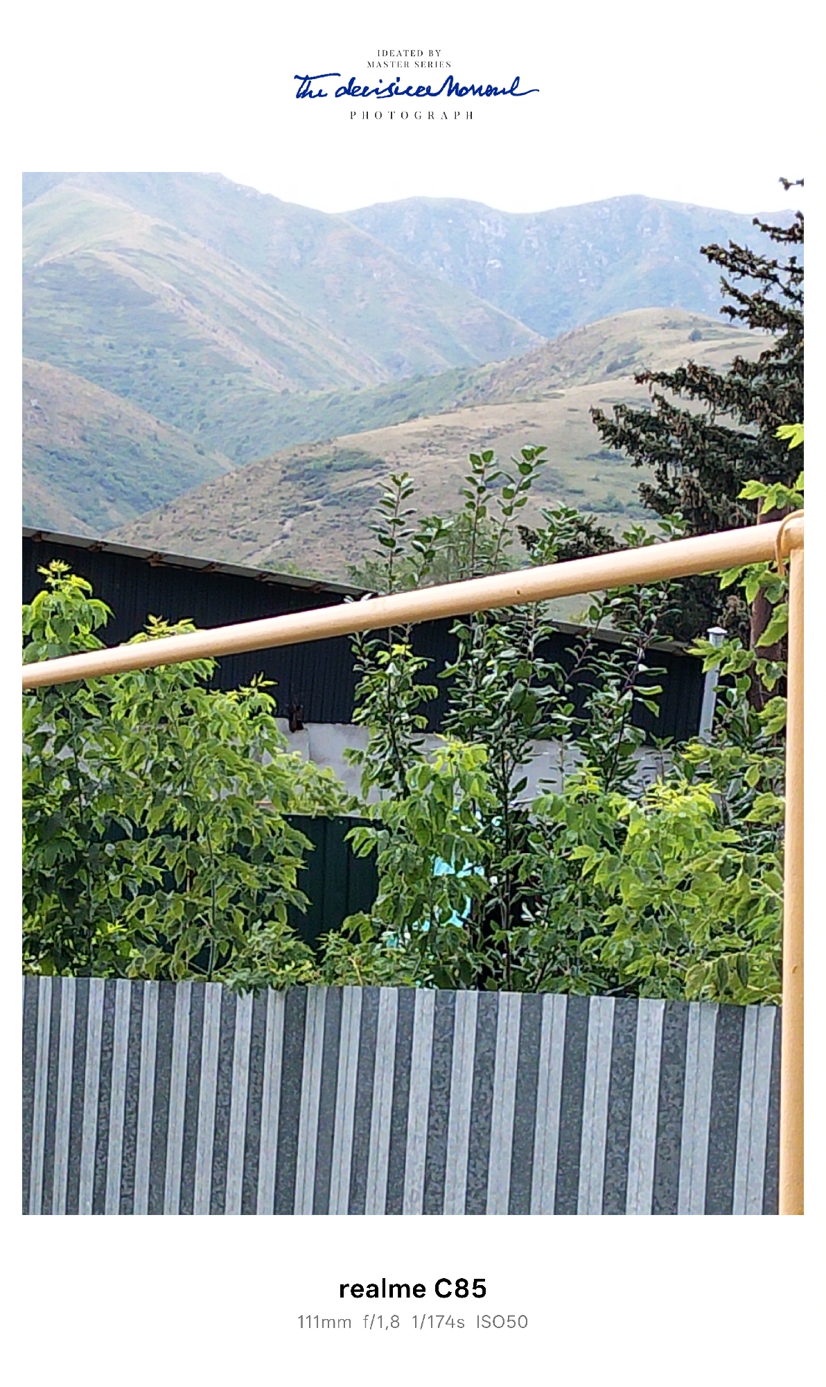 The image is a photograph with a mountain landscape in the background and a residential area in the foreground. In the upper portion of the image, there is a watermark with text that reads "IDEATED BY MASTER SERIES The decisionl Monoul PHOTOGRAPH". The mountain range is characterized by dry, grassy slopes and rocky outcrops, illuminated by natural light, suggesting it is daytime.

In the middle ground, a dark, corrugated metal roof of a building is visible, partially obscured by lush green foliage of trees and bushes. A horizontal, light-colored pipe or railing runs across the middle of the image, in front of the foliage and the building.

The foreground features a vertical, ribbed fence made of corrugated metal, with alternating dark and light gray stripes. Some green foliage is visible above the fence. The bottom of the image contains text overlay indicating camera specifications: "realme C85 111mm f/1,8 1/174s ISO50".

The overall scene depicts a rural or semi-rural setting, likely within or on the outskirts of the city of Tekeli, Kazakhstan, given the characteristic mountainous terrain. The image captures a view from a residential area looking towards the mountains. There are no people or animals visible in the photograph.