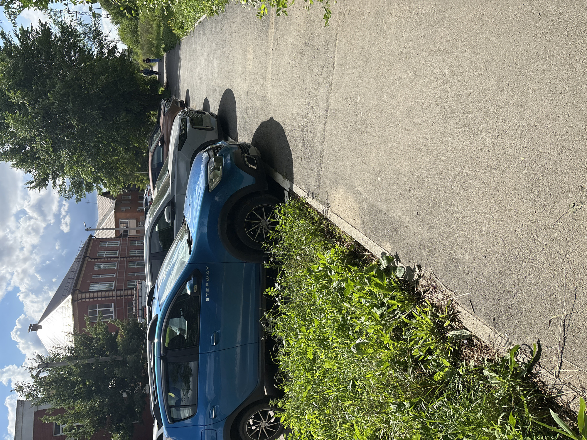 The image depicts a street scene in Orsk, Russia, on a sunny day. In the foreground, lush green grass and weeds grow along the edge of a paved walkway. Several cars are parked in a row on the other side of the walkway, including a blue Dacia Sandero Stepway. Behind the cars, a brick building with white-trimmed windows and a peaked roof stands under a partly cloudy sky. Further down the street, a large tree provides shade, and two individuals, a woman and a child, are walking away from the camera. Power lines are visible in the background.