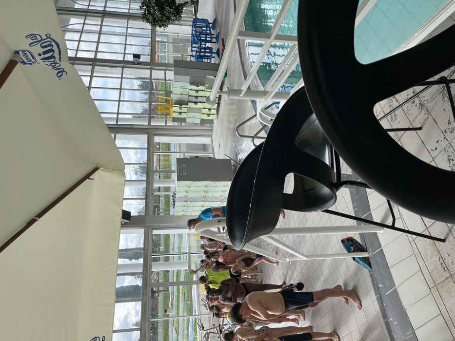 This image depicts an indoor swimming pool facility, specifically the Piscine Antinéa in Andrézieux-Bouthéon, France, identified by the "Nestlé AntínéA" logo on an overhead cream-colored canopy. Approximately 20-25 children, primarily of elementary school age and dressed in swimwear, are gathered on the white and grey tiled pool deck, forming a line. An adult male in a blue shirt is positioned among them, appearing to supervise. In the immediate foreground, two black, high-backed bar stools partially obstruct the view. The background features large, arched floor-to-ceiling windows looking out onto an exterior landscape with trees, green space, and an overcast sky. Inside, along the windowed wall, are several stacks of light green lockers and a grey locker unit displaying a sign that reads "INFORMATION A VENIR". An inverted yellow chair rests on top of one locker stack. To the right, a blue plastic barrier is visible near the pool's edge. The pool itself contains clear, light blue water, bordered by a white railing. A blue kickboard is visible near a floor drain on the deck.