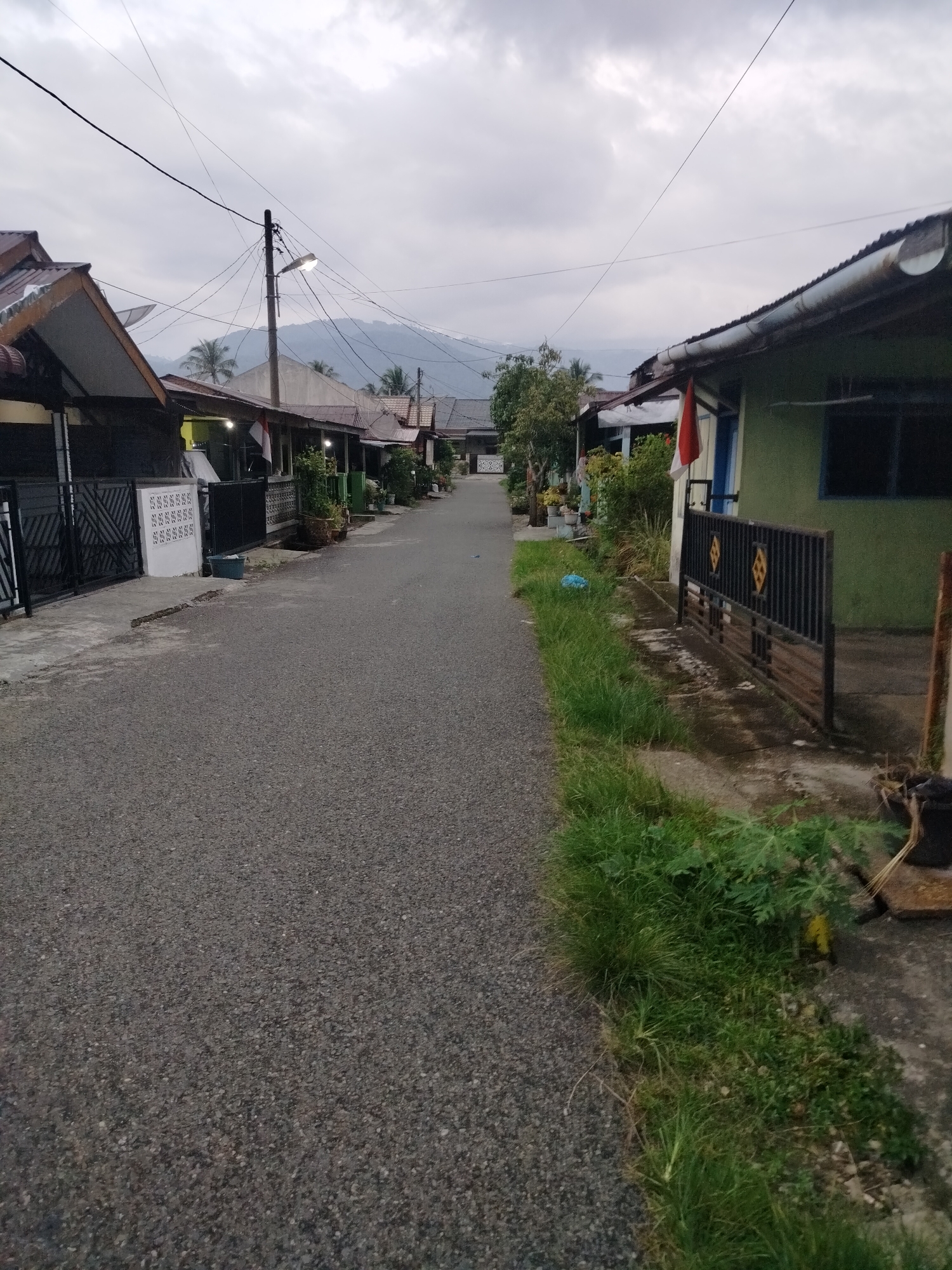 The image shows a residential street in Kumbangindah, Indonesia, under an overcast sky. The asphalt road runs through the center of the frame, lined on both sides by single-story houses with corrugated metal roofs. The houses have various fence styles, some with decorative ironwork and others with solid white walls. Potted plants and some greenery are visible along the sides of the road and in front of the houses. A utility pole with numerous wires is prominently positioned on the left side of the street, and a street lamp is illuminated at the top of the pole. In the background, mountains are faintly visible through the hazy sky. There are no people visible in the image.