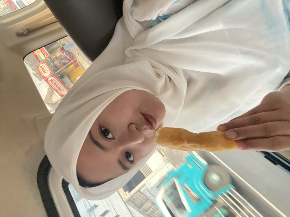A woman in a white hijab sits inside a car while eating. Her face is turned towards the camera, with her eyes looking directly at the viewer. The background shows an outdoor scene, visible through the car windows, including signs and buildings. She is holding a long, golden-brown pastry in her hand, taking a bite. The car's interior and other details of the outside environment are also visible in the frame.