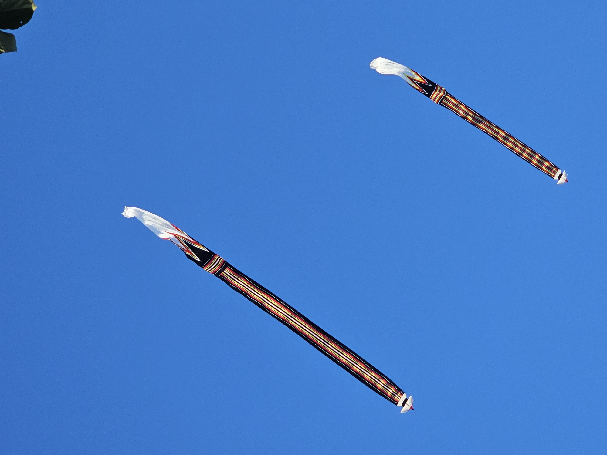 Two traditional Balinese kites, known as "layangan" or "ogoh-ogoh," are flying against a clear blue sky in Denpasar, Indonesia. The kites are long and slender, with a central pole structure. They are decorated with colorful woven patterns featuring black, white, red, and yellow stripes, creating a vibrant, geometric design. The top of each kite is adorned with a white, streamer-like material that trails in the wind. The kite in the upper left is larger and positioned diagonally, while the smaller kite is in the lower right, also at an angle. The scene is set outdoors, with a hint of green foliage visible in the top right corner, suggesting a natural or park-like environment within the city.