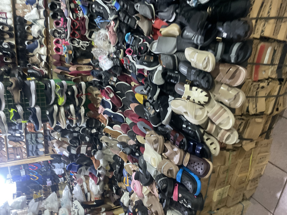 This image depicts a densely packed retail shoe display within an indoor market stall or shop located in Nganguk, Indonesia. The scene is dominated by a vast assortment of new and used footwear of various styles, sizes, and colors, including sneakers, sandals, flip-flops, slip-ons, and a few closed-toe shoes. The shoes are arranged in a disorganized yet functional manner, with many items piled directly on top of each other on multiple levels.

In the foreground and midground, a wide variety of open-toed sandals and clogs, some resembling Crocs-style footwear, are visible. Colors range from black, white, and various shades of brown and beige to brighter hues like pink and blue. Many shoes appear to be made of synthetic materials, rubber, or plastic.

The background shows more shoes displayed on shelves or hanging racks, with numerous pairs stacked on elevated platforms. Some of these shoes include athletic sneakers, dress shoes, and more casual footwear. The display structure consists of rudimentary wooden shelves or crates, and below the primary display, stacks of cardboard boxes, likely containing additional inventory, are visible. The lighting is artificial, suggesting an indoor setting. No individuals are present, and no actions or interactions are depicted. The overall impression is that of a bustling, high-volume shoe vendor.