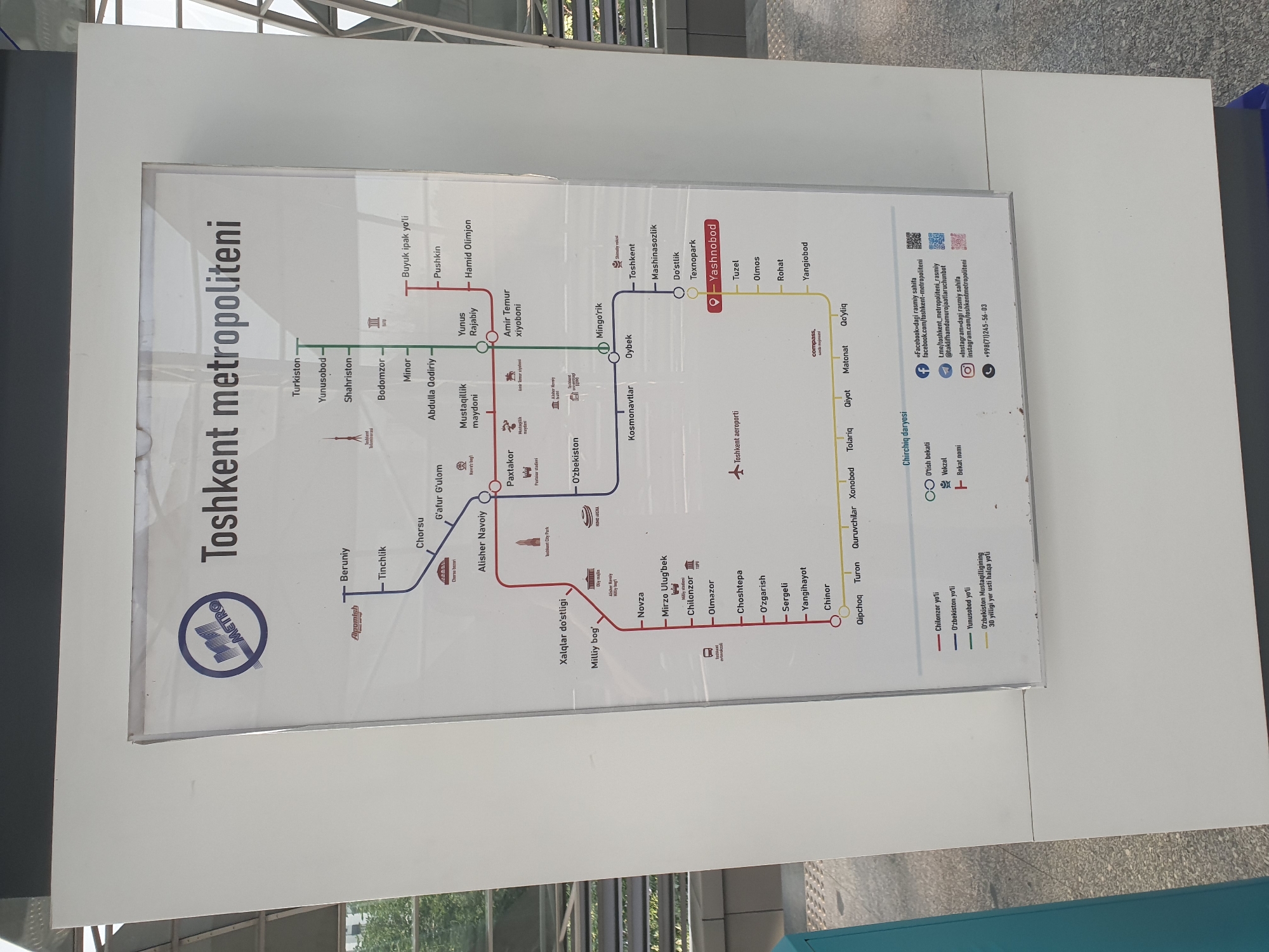 The image shows a detailed map of the Tashkent Metro system. The map is mounted on a white panel and appears to be displayed in a public area, likely a metro station, given the presence of a blue and silver metro logo and the overall design. The map itself is colorful, with different lines represented by distinct colors. Various station names are clearly labeled in what appears to be Cyrillic and Latin scripts. Icons are used to denote landmarks or specific features associated with certain stations, such as the Tashkent TV Tower, stadiums, and cultural buildings. The map also includes a legend explaining the different colored lines and symbols. Towards the bottom right of the map, there are contact details for the Tashkent Metro, including social media icons for Facebook, Telegram, and Instagram, as well as a phone number. The environment around the display is neutral, with a hint of a modern urban setting, possibly an indoor transit hub. The overall impression is an informative and well-presented public transportation map.