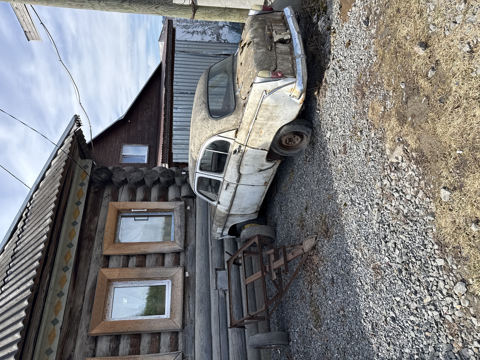 The scene depicts a dilapidated white car, likely a vintage model, parked next to a traditional wooden log cabin structure in Tyulyuk, Russia. The car is heavily weathered and appears abandoned. The cabin features three framed windows with modern plastic frames inset into the wooden logs, indicating some level of renovation. To the right of the car, the ground is covered in gravel and sparse dry grass. Behind the car is a corrugated metal structure. In the background, a brown, gable-roofed building is visible under a sky with scattered clouds. A rusted trailer is adjacent to the car, adding to the impression of disuse and abandonment.
