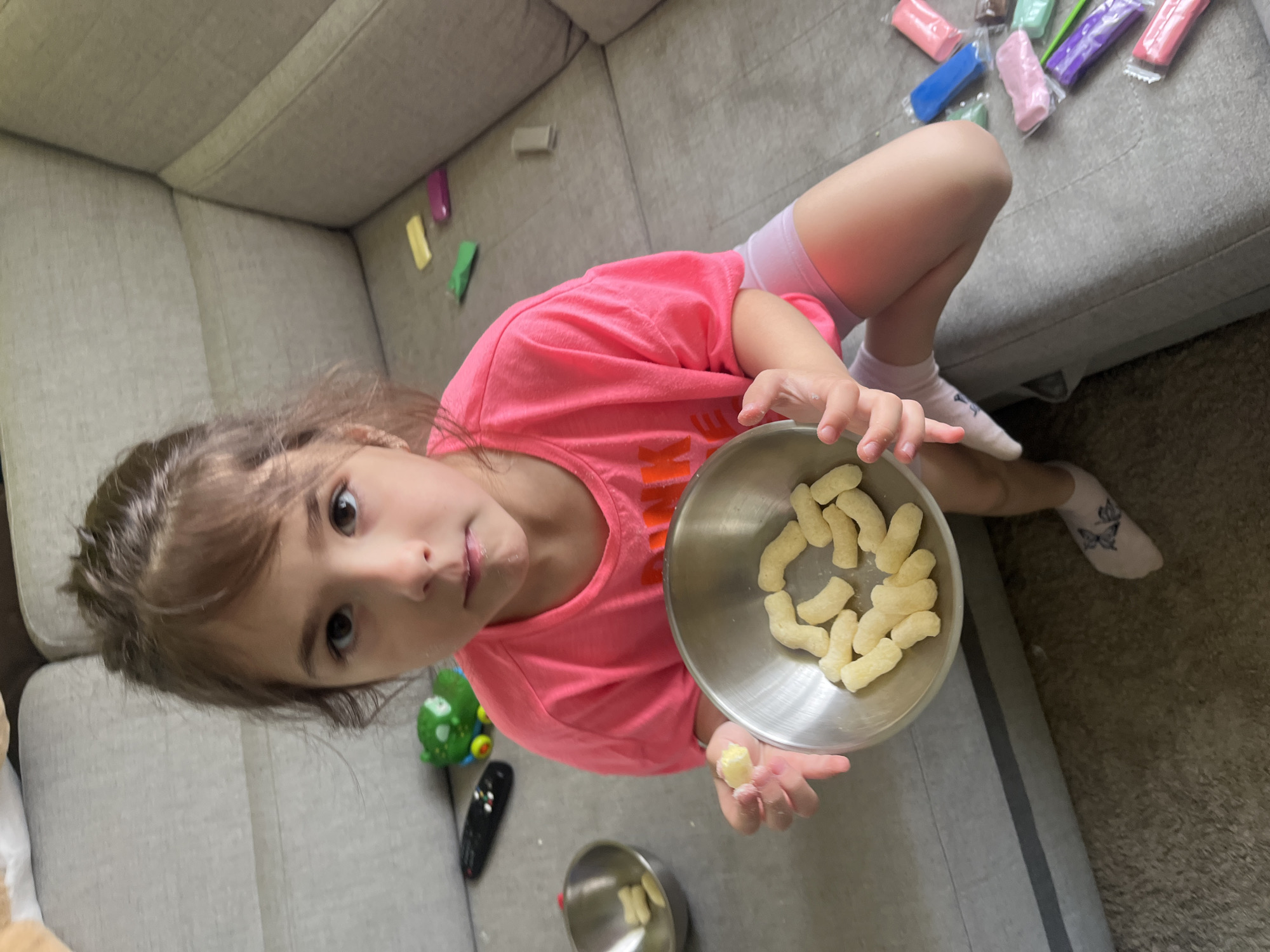 A young girl with brown hair pulled back sits on a grey sofa in what appears to be a living room. She is wearing a bright pink t-shirt and light pink shorts. In her hands, she holds a small metal bowl filled with puffed corn snacks, and she is holding one snack up as if to eat it. To her left, another metal bowl with snacks is visible. Various colored sticks of modeling clay are scattered on the sofa near her legs, along with a small green toy and a black remote control. The overall environment suggests a casual, indoor setting.