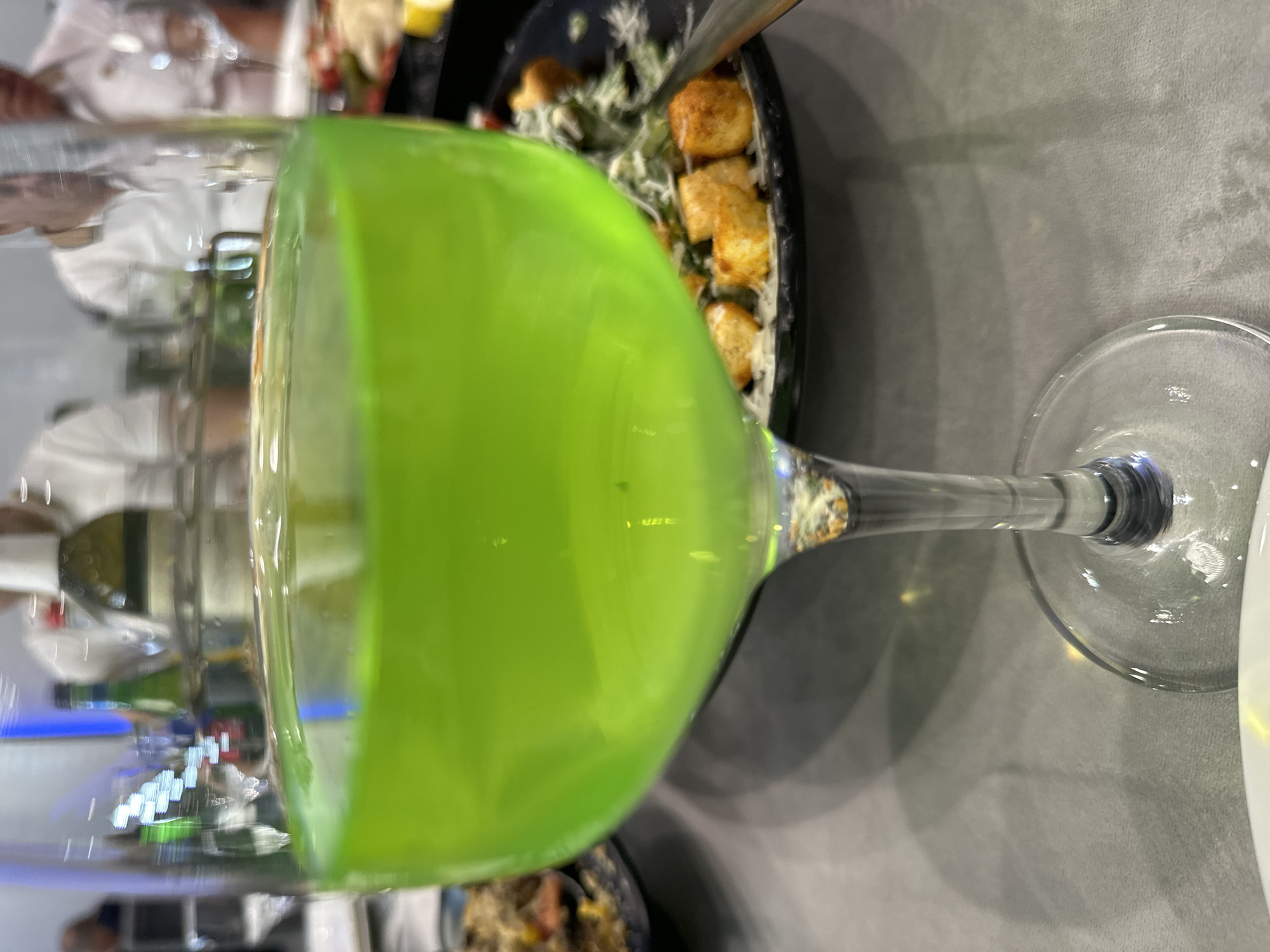 The image depicts a close-up view of a wine glass filled with a bright green liquid. The glass is positioned centrally on a gray, textured surface, likely a tablecloth. In the background, slightly out of focus, are several people and plates of food, suggesting a dining setting. To the left, a plate with a dark rim holds what appears to be a salad with croutons and greens. Further back, a person in a white shirt is visible, and behind them, another person wearing a light-colored polo shirt can be seen. The lighting in the scene is artificial and appears to come from overhead or the side, casting soft shadows. The overall atmosphere is casual and social.