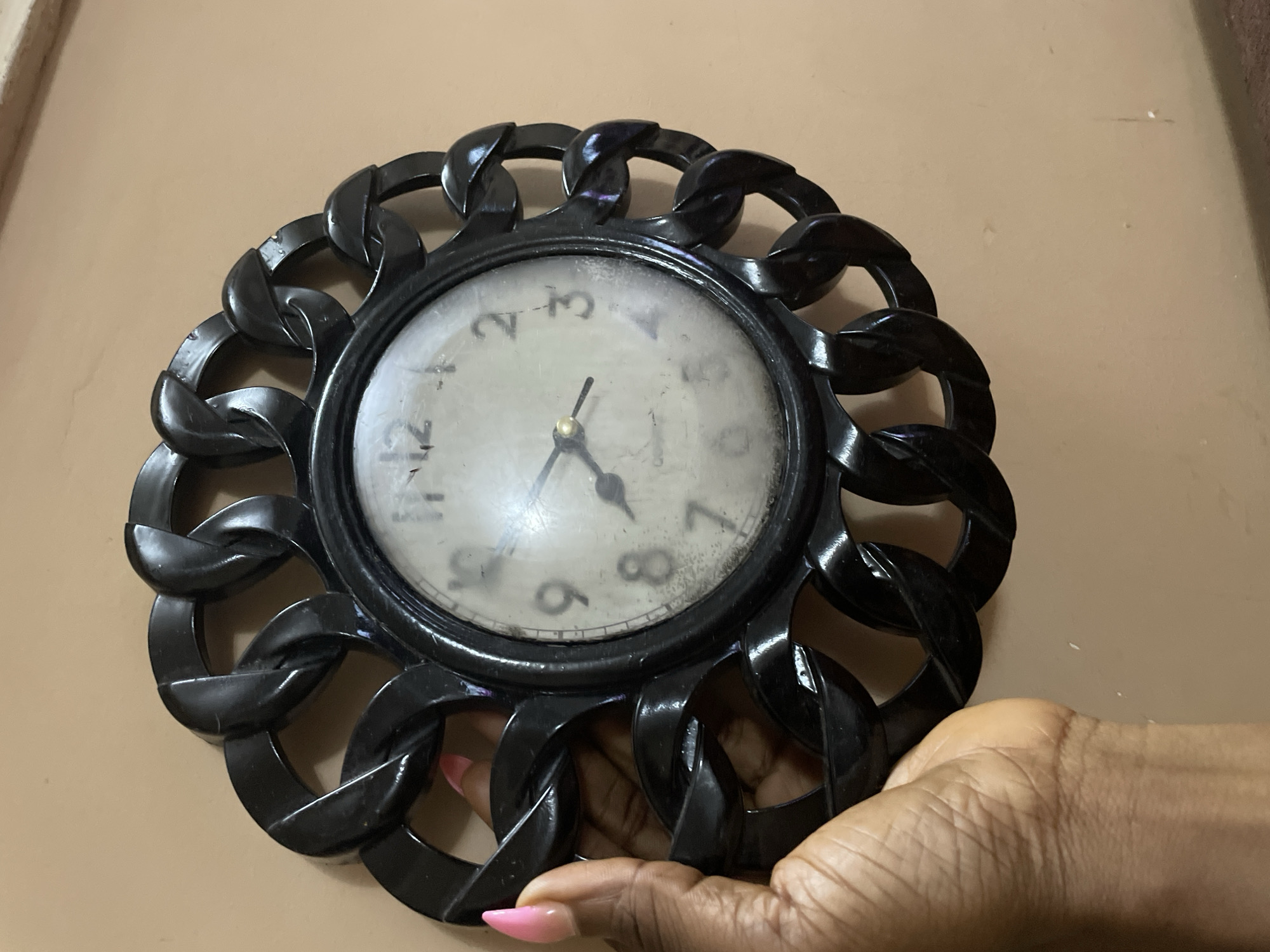 The scene depicts a close-up, angled view of a dark-skinned hand with bright pink, pointed artificial nails, interacting with a circular wall clock mounted on a plain, light brown wall. The location is specified as the city of Narayi, Nigeria. The clock features a black, ornate frame designed with interconnected, dark chain-like links forming its perimeter. The central clock face is light-colored, possibly off-white, and appears significantly dusty or soiled, with visible smudges and imperfections across its surface and possibly on its protective cover. Arabic numerals are used for the hours, with numbers such as 2, 3, 4, 7, 8, 9, 12, and 1 partially visible through the grime. Two black clock hands are visible, indicating an approximate time of 7:05. The word "Quartz" is faintly discernible on the lower half of the clock face. The hand is gripping or steadying the left side of the clock's ornate frame, with the thumb and index finger visible. The wall behind the clock is unadorned, exhibiting minor surface variations and specks, suggesting an indoor environment.