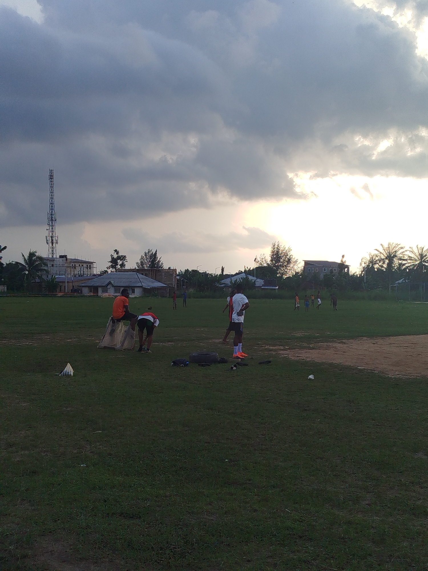 The image shows a grassy field in Ikot Mbonde, Nigeria, under an overcast sky. In the foreground, two people are near what appears to be a sack; one person is sitting on it, and the other is bending over. Nearby are black tires and other objects on the ground. In the midground, several people are playing or exercising on the field. Buildings and vegetation, including palm trees, are visible in the background, along with a tall communication tower. The time of day appears to be late afternoon or early evening, suggested by the lighting through the clouds.