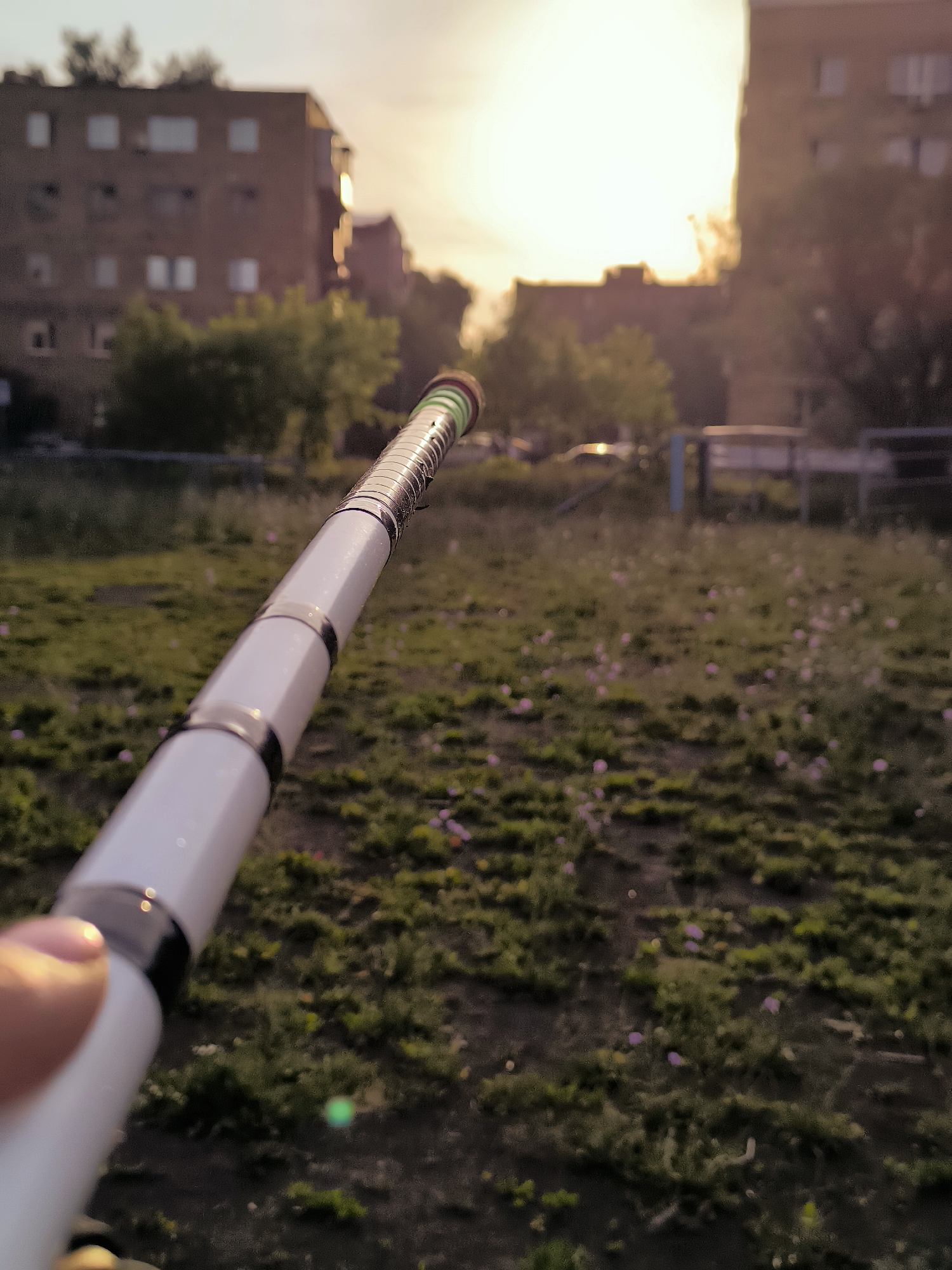 A close-up, shallow depth-of-field shot shows a person's thumb and forefinger holding a white electronic cigarette device with metallic accents. The device is positioned diagonally across the frame, extending from the bottom left corner towards the upper right. The background is blurred, featuring a grassy field with scattered small purple flowers. In the mid-ground, the outlines of residential buildings are visible, suggesting an urban setting. The sky above is bright, indicative of sunset or sunrise, with the sun's glare creating a hazy glow. The scene appears to be an outdoor recreational area, possibly a park or a courtyard within a residential complex in Qarağandı city administration, Kazakhstan.