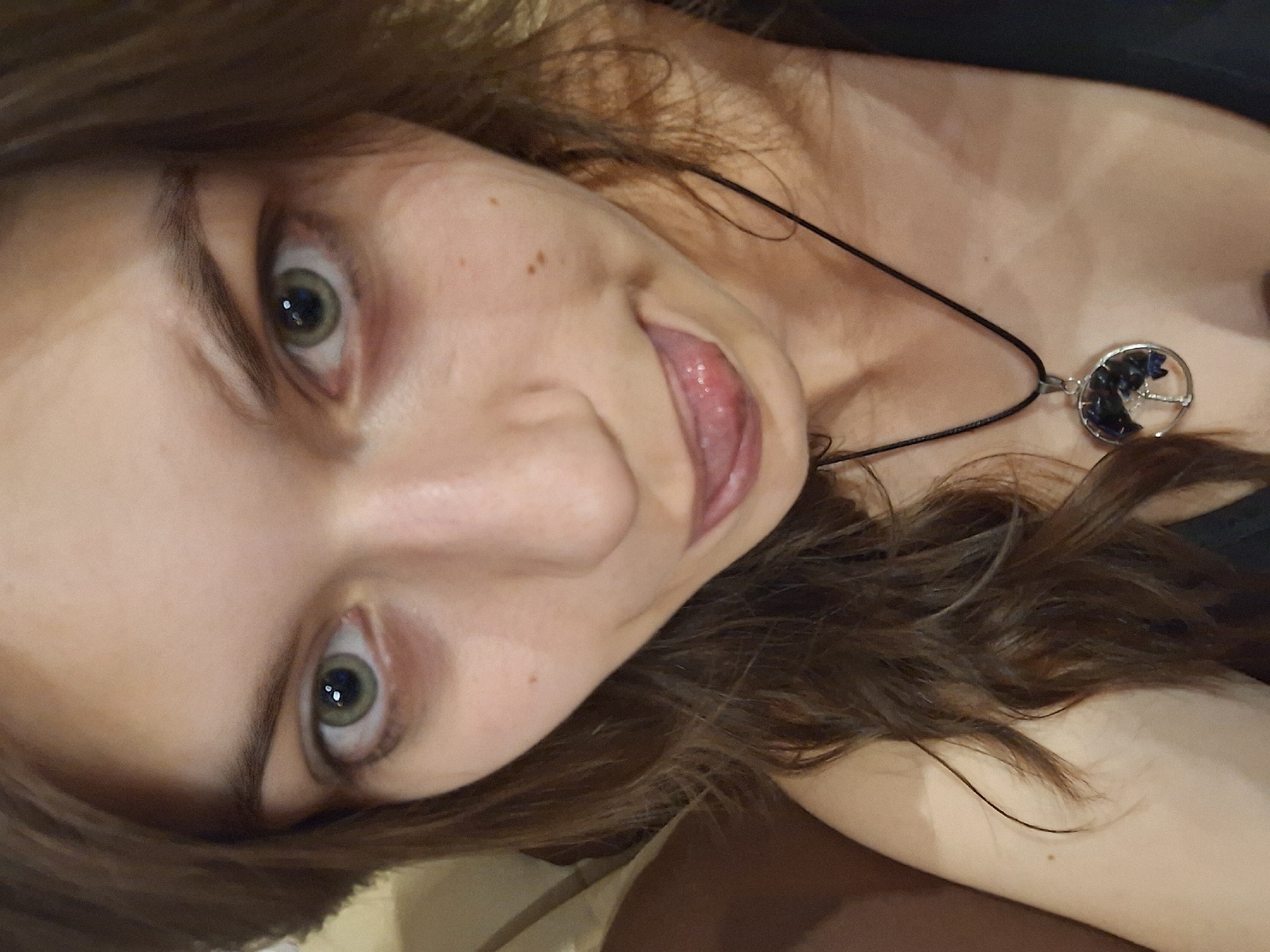 A selfie of a fair-skinned person with light brown hair and green eyes sticking out their tongue. The person is wearing a black cord necklace with a circular pendant filled with dark blue and clear stones. The background is out of focus. The location is not discernable from the image.