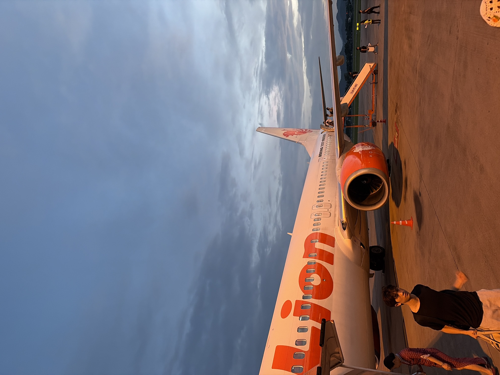 An airplane with "Lion" written in large orange letters on its side is parked on an tarmac at an airport in Suthep Town Municipality, Thailand. The plane is a Boeing 737-900ER, indicated by text on its tail. People are boarding or disembarking the aircraft via a set of stairs. A man in a black t-shirt and light-colored shorts is walking in the foreground, carrying a bag. A woman in a patterned uniform stands to the left of the frame. The sky is cloudy with hints of sunset colors. The overall scene depicts a typical airport environment during dusk.