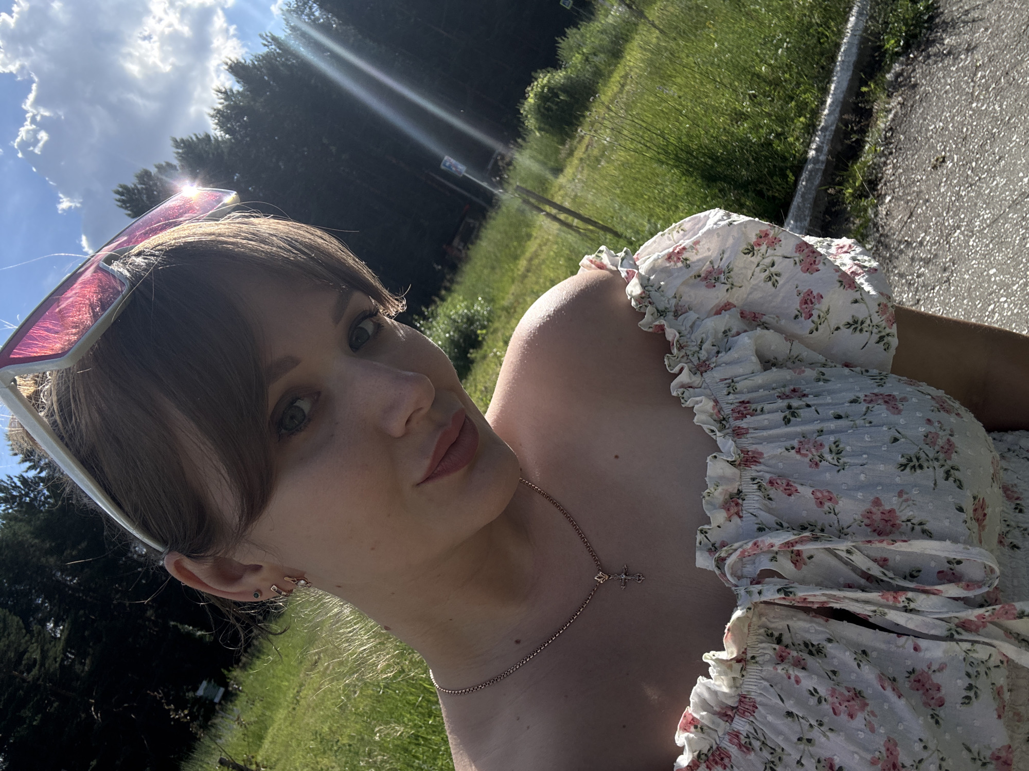 In a sun-drenched outdoor scene, a young woman is captured in a selfie. She is wearing a light-colored, floral-print dress with ruffled sleeves and a necklace featuring a cross pendant. Large, pink-tinted sunglasses are perched on her head. Her hair is brown and styled with bangs, and she has visible earrings and piercings in her ear. The background consists of lush green grass, tall trees, and a bright blue sky with scattered clouds, suggesting a pleasant day. The sun is shining brightly, casting lens flares across the image and indicating it is likely daytime. The woman's expression is relaxed and she is looking towards the camera. The scene appears to be a natural or park-like setting, possibly near a paved path visible on the right side of the image. No visible text is present in the image.