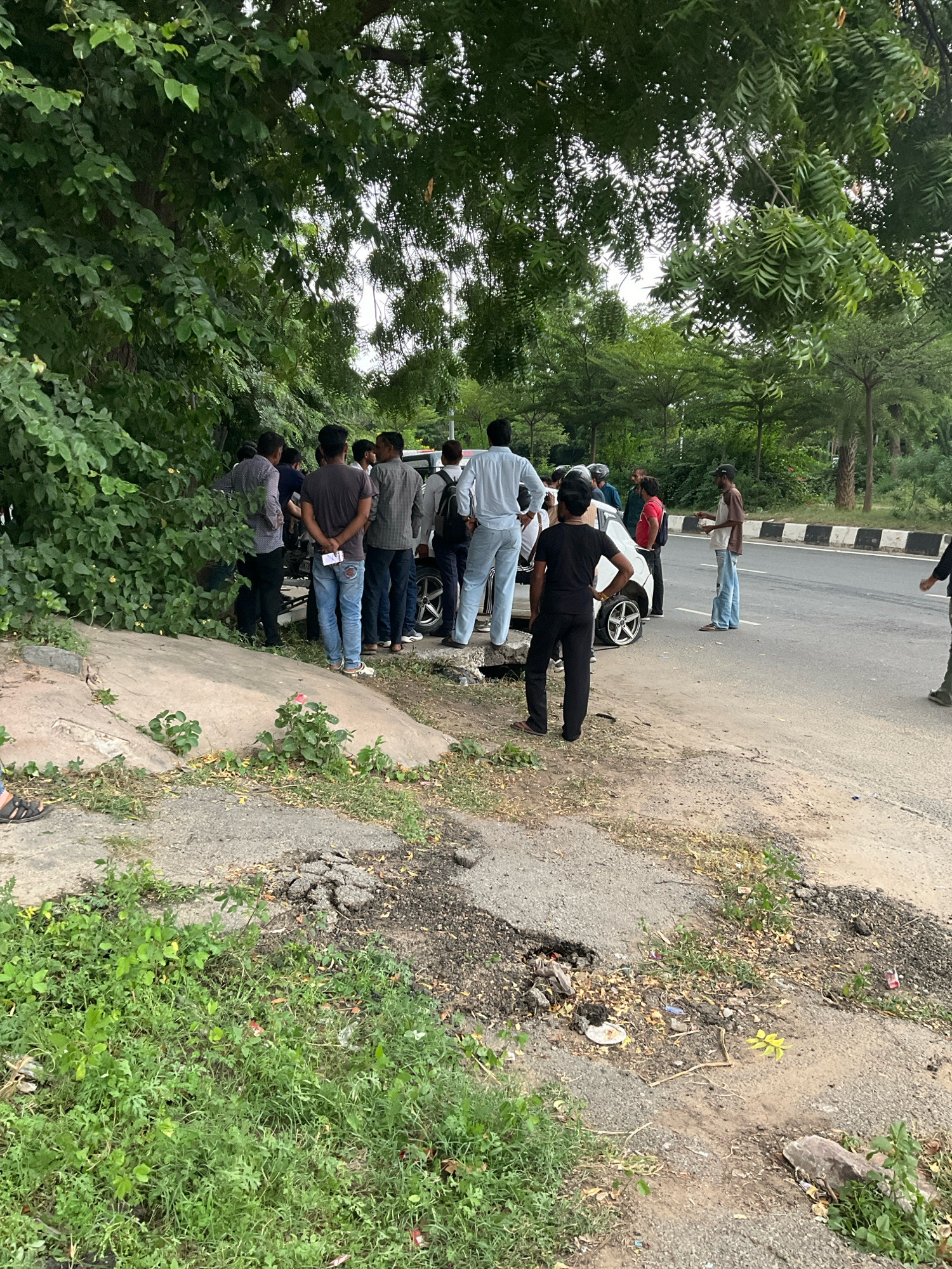 An overhead perspective captures a group of individuals gathered around a white car positioned on the side of a road, partially off the pavement and on an incline with lush green foliage. The scene suggests a roadside incident, possibly a breakdown or an accident, as the car's front passenger side wheel appears to be turned at an unnatural angle.
Several men are standing near the car, some looking at it, while others are in conversation. One man in a light blue shirt and light-colored pants stands with his back to the camera, appearing to observe the situation. To his left, a man in a dark t-shirt and ripped blue jeans stands with his hands behind his back, facing away from the car and towards the left of the frame. Further left, a cluster of men are gathered closer to the car, their interaction unclear.
Across the road, a black and white striped barrier runs along the edge of the pavement, indicating the car is on the shoulder of a paved road. The environment is characterized by dense green trees and foliage, suggesting a location with abundant vegetation. The ground surface is a mix of cracked asphalt, dirt, and scattered debris, including some small rocks and dried leaves.
The sky is overcast, with diffused light suggesting it is daytime, likely in a tropical or subtropical climate given the greenery. The presence of a paved road, striped barriers, and the general urban landscaping point towards a location within the Jaipur Municipal Corporation, India, consistent with typical roadside environments in the region. No specific landmarks or identifying signs are visible to pinpoint the exact location within the corporation.