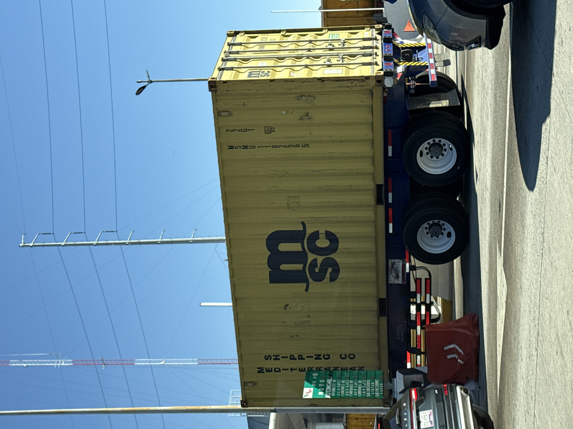 This image captures an outdoor scene on a paved road in Santa María Moyotzingo, Mexico, under a clear blue sky with bright sunlight. The central subject is a large, yellow corrugated shipping container, prominently displaying the black "m sc" logo of MSC (Mediterranean Shipping Company) on its side. Vertical text reads "MEDITERRANEAN SHIPPING CO." Another visible side of the container is marked "MSMU 1182565" and "22G1," along with a "tlc 83" symbol. The container rests on a blue tandem-axle trailer, which features white multi-spoke wheels and red and white reflective safety striping along its lower edge.
In the foreground to the left, the rear section of a dark-colored SUV is visible, displaying a Mexican license plate partially reading "XTC-579-D." Adjacent to this vehicle and the trailer is a red plastic road barrier marked with white chevron symbols. To the right, the front portion of another dark-colored vehicle is partially visible.
The background features a tall, red and white striped lattice communications tower on the left, with power lines or communication cables spanning the sky. Another tall, thin pole, possibly a utility pole, is also visible. On the right, a utility pole with a street light fixture is present. A green rectangular sign, partially obscured, is attached to a pole near the front of the container, displaying text in Spanish that appears to categorize vehicles (e.g., "AUTOMOVIL," "CAMIONES," "CARGA"). Strong shadows are cast on the light-colored paved road surface, indicating the sun's position. No individuals are visible in the scene.