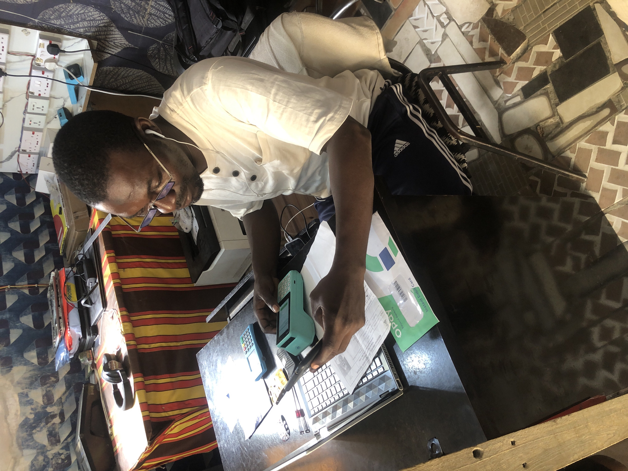The image depicts a man sitting at a desk in what appears to be a small shop or office in Ado, Nigeria. The man, wearing glasses and a white short-sleeved shirt, is looking down at a laptop and a payment terminal, with a pen in his right hand. A smartphone is resting on the laptop. On the desk, there is also a blue calculator and some documents, including what looks like a debit card envelope. Behind the desk, a colorful striped cloth covers the wall or a partition. Electrical outlets are visible on the wall to the right, with some devices plugged in. The floor is tiled with a mosaic pattern. The overall impression is that of a business transaction or administrative work being conducted in a modest urban setting.