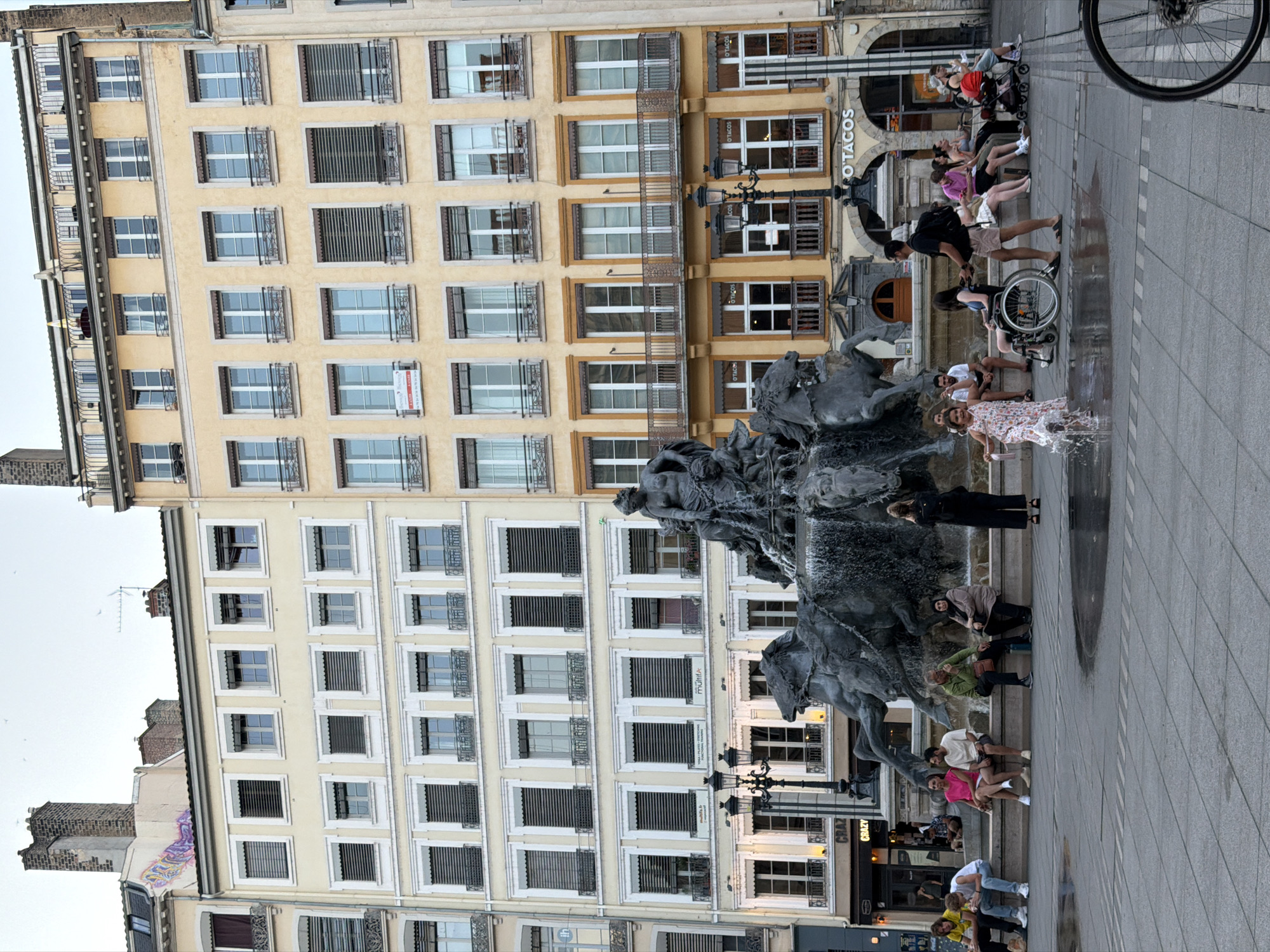 The image shows a view of Place Louis Pradel in Lyon, France, with the Théâtre des Célestins on the right and the Hôtel de Ville in the background. A large, ornate fountain with sculptures of horses and figures is in the foreground, with water spraying from it. Several people are sitting on the edge of the fountain or standing nearby. A young girl in a floral dress is running through the water. The buildings in the background are typical of Lyon, with their light-colored facades and numerous windows. The sky is overcast, and the lighting suggests it is late afternoon or early evening.