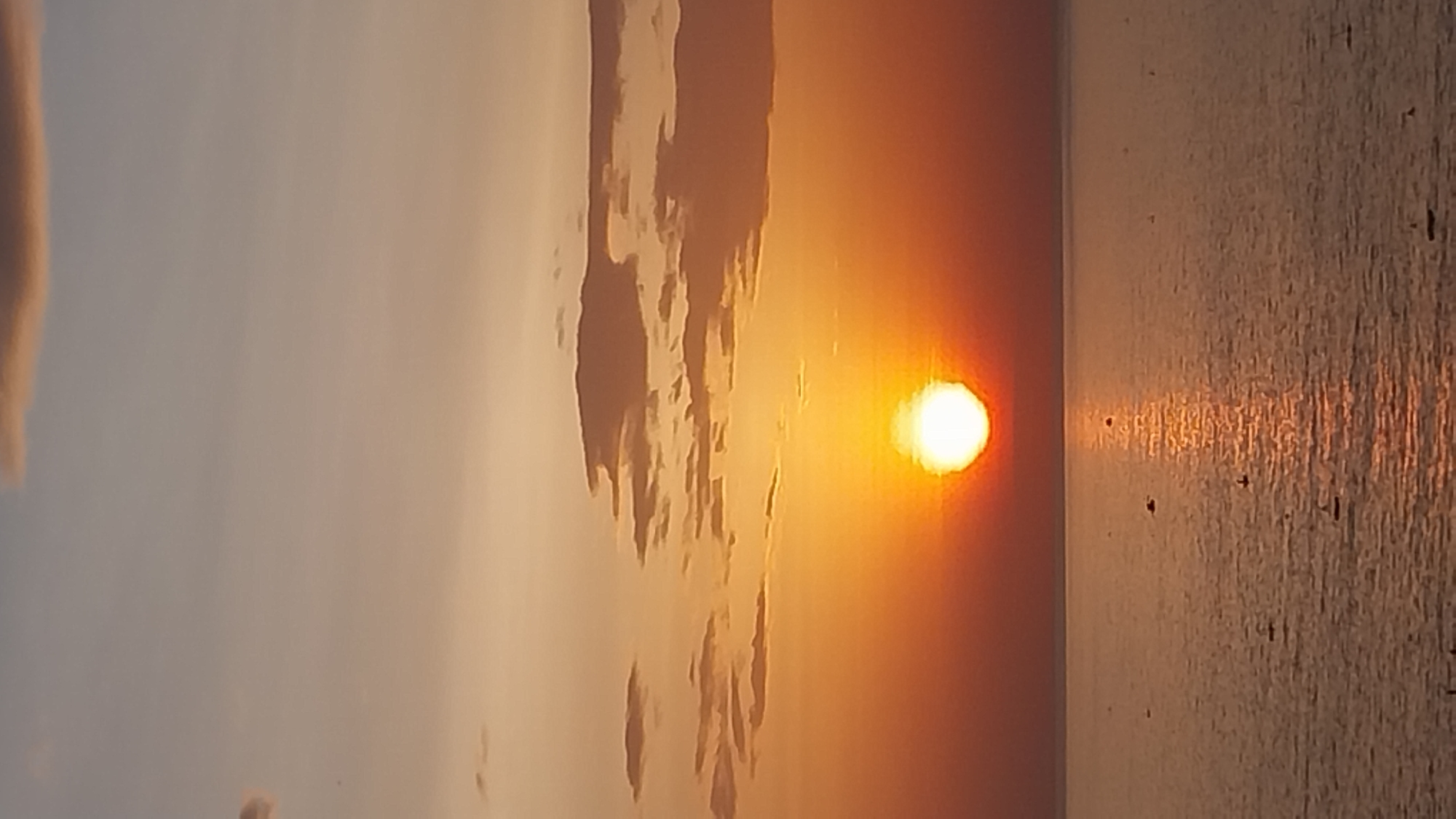 A vertical, medium shot captures a tranquil sunset over a calm body of water. The sun, a bright orb, is positioned in the center of the frame, casting a golden glow that saturates the sky with hues of orange and peach. Wispy clouds, tinged with the same warm colors, are scattered across the upper portion of the image. The water below reflects the vibrant sky, creating a shimmering pathway of light extending from the sun to the foreground. Several small boats are visible on the water, their dark silhouettes contrasting with the bright reflection. The horizon line is sharp and distinct, separating the sky from the sea. The overall atmosphere is serene and peaceful, indicative of a quiet evening in a coastal area, potentially in Tuy Phong 4, Vietnam, known for its beautiful sunsets.