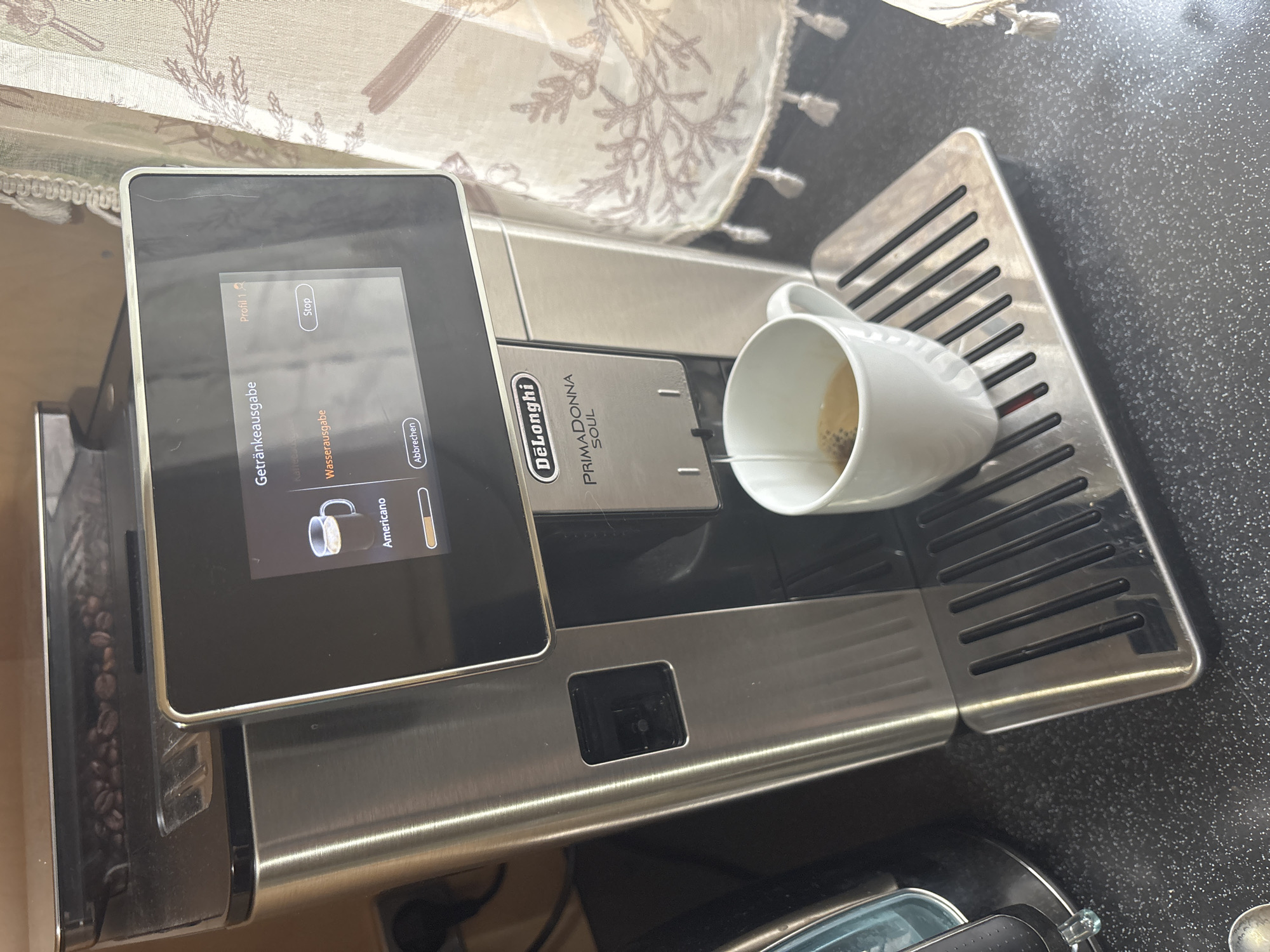 The image shows a close-up of a DeLonghi PrimaDonna Soul coffee machine dispensing an Americano into a white mug. The coffee machine has a stainless steel finish and a digital display showing the beverage selection screen. Coffee beans are visible in the hopper at the top of the machine. The mug is positioned on the drip tray, and a stream of coffee is pouring into it. The background features a patterned curtain and a speckled countertop. The setting appears to be a domestic kitchen.